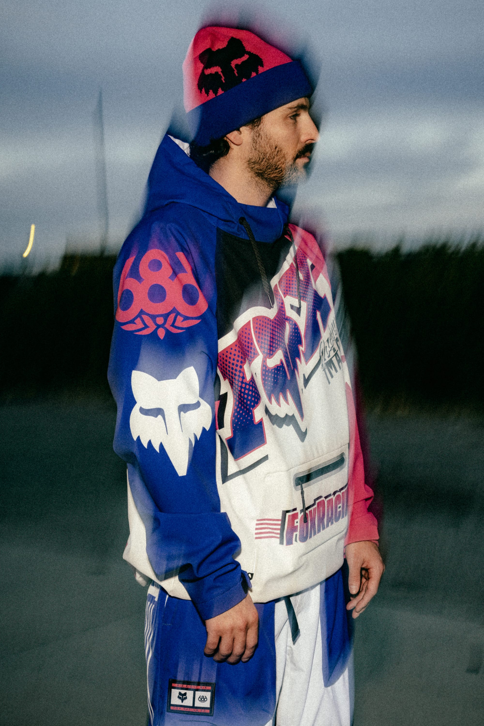 A man wearing the 686 Men's Fox Racing Waterproof Hoody by 686, featuring bold pink, blue, and white graphics with large logos, stands outdoors at dusk. The scene has a blurred, double-exposure effect.