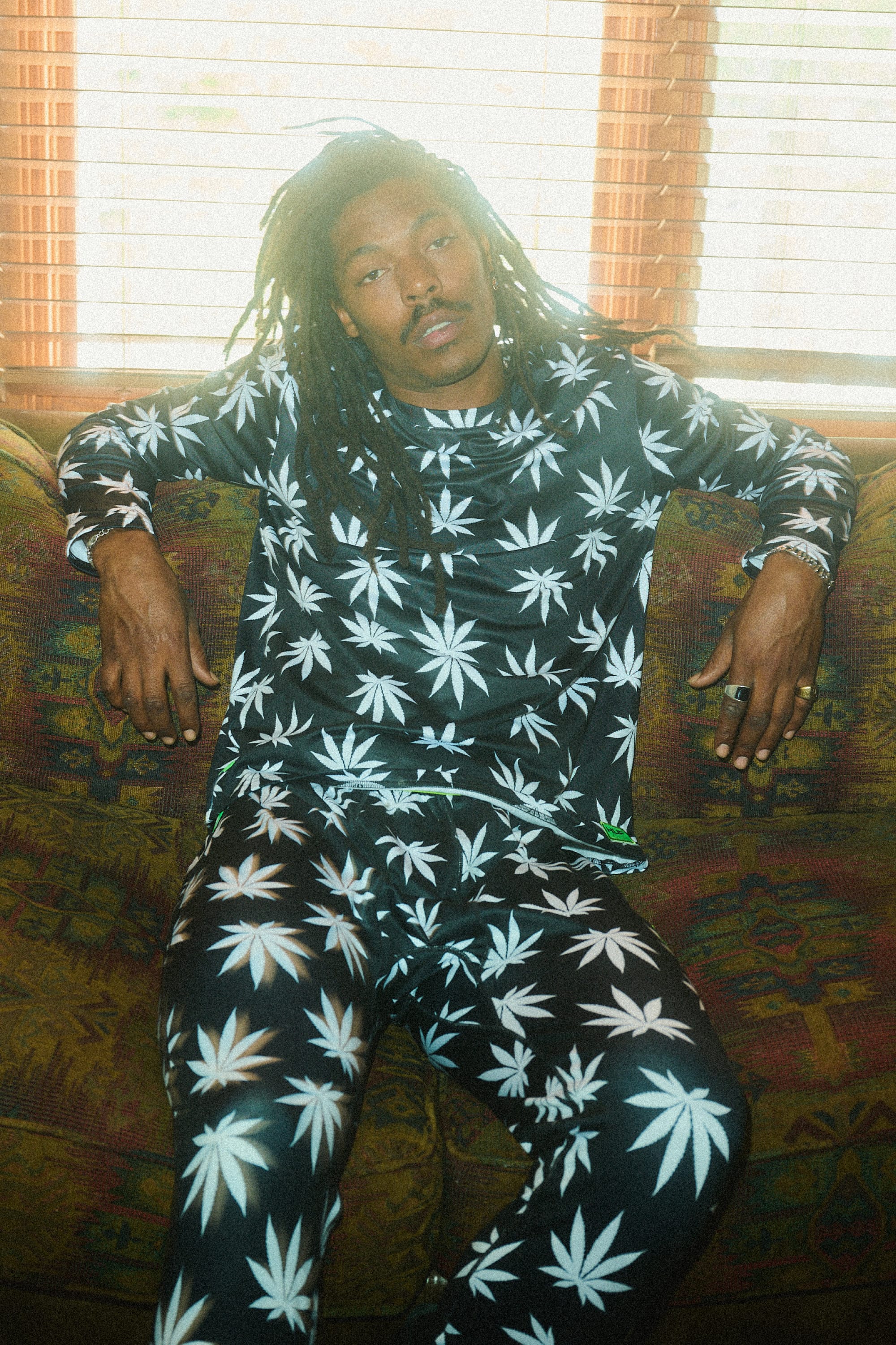 A man with long dreadlocks relaxes on a couch, wearing the 686 Mens Plantlife Base Layer Bottom by 686, featuring bold white marijuana leaf patterns on black polyester spandex as sunlight streams through the blinds.