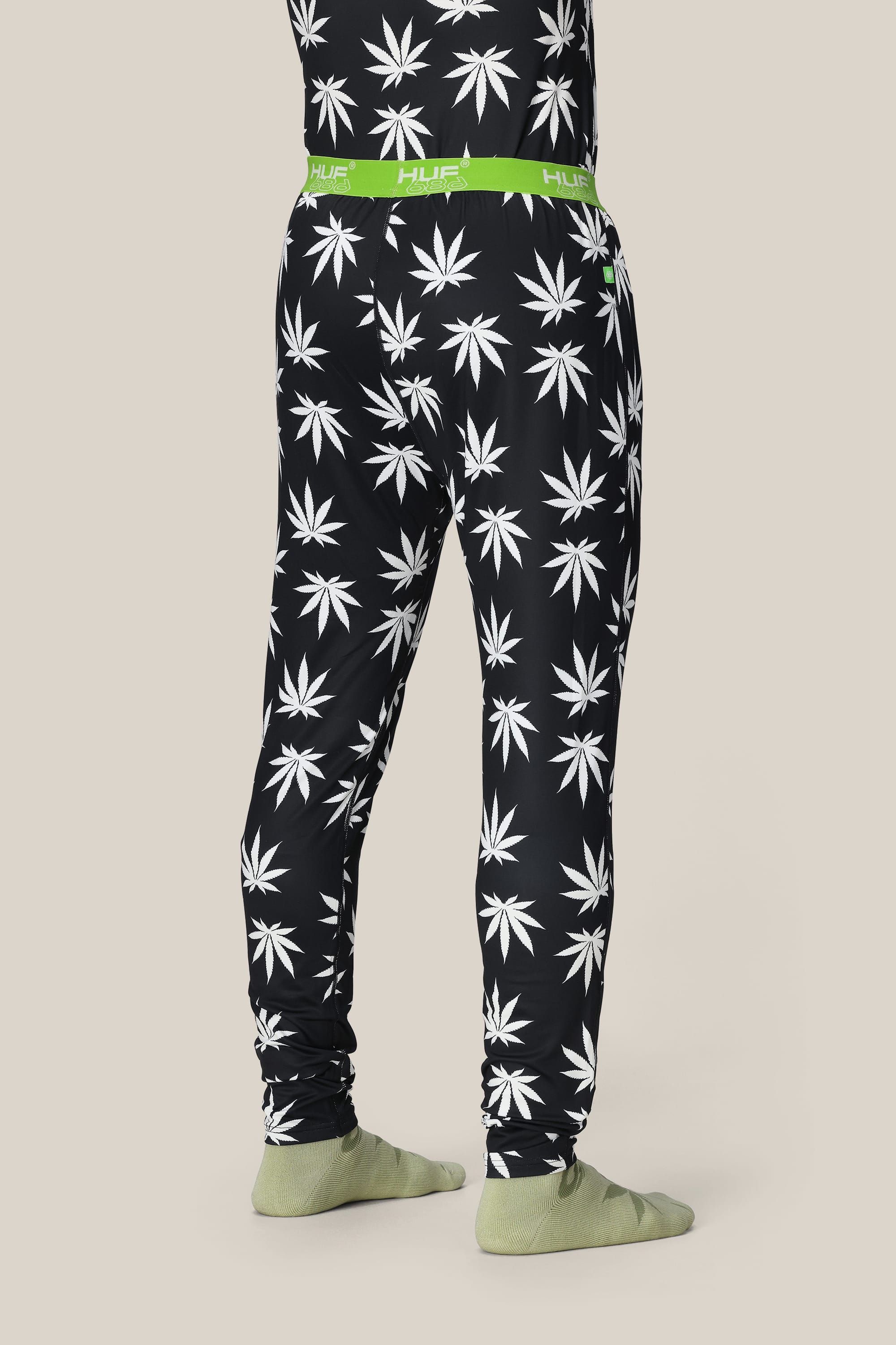 A person stands facing away, wearing 686 Mens Plantlife Base Layer Bottom by 686 with a white cannabis leaf pattern, green waistband labeled HUF, and light green socks, set against a beige background.
