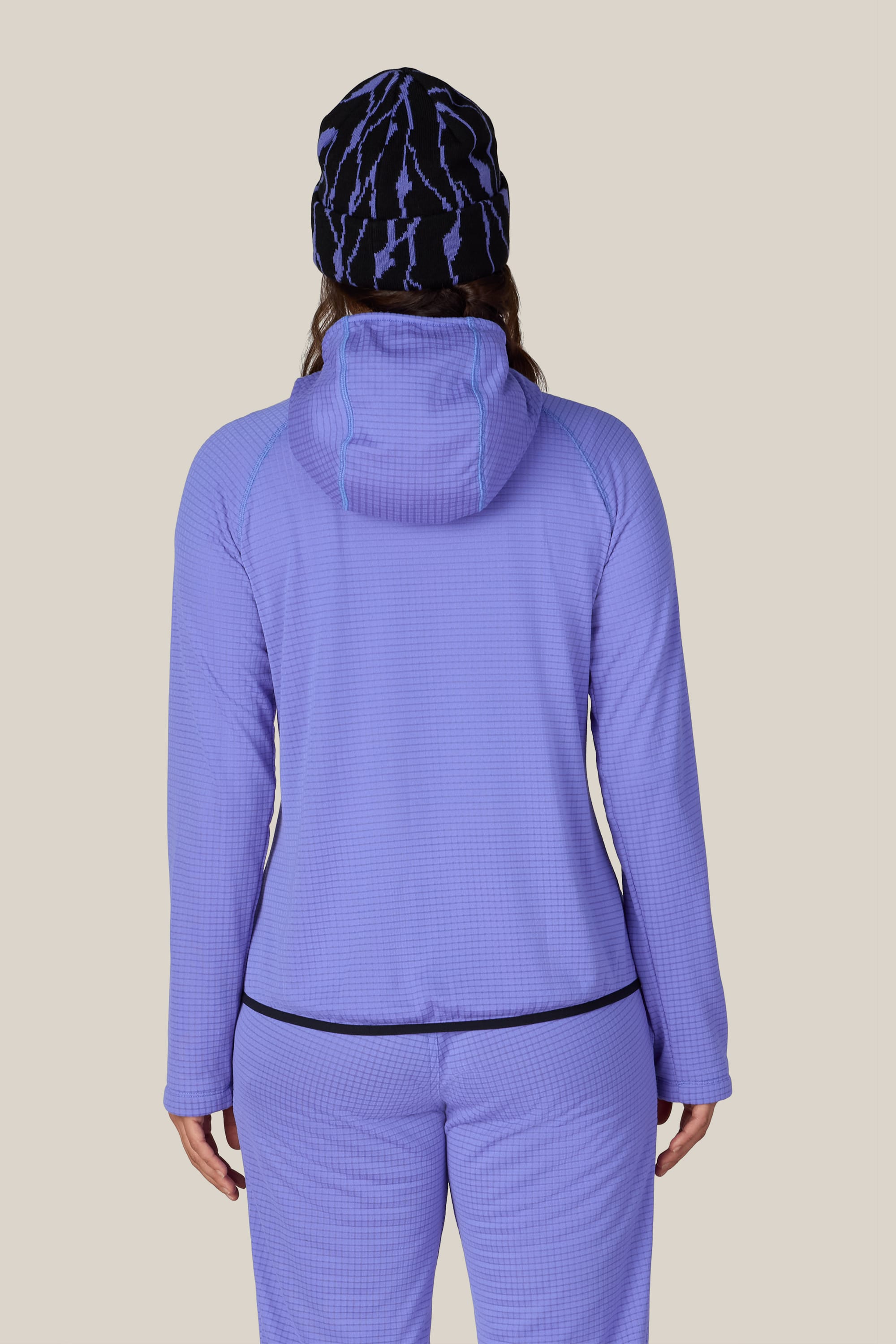 A person with long hair, seen from behind, wears the 686 Women’s Mission Grid™ Hoody in purple textured fabric paired with matching pants and a black-and-blue patterned beanie. The stylish comfort of the 686 ensemble stands out against the plain beige background.