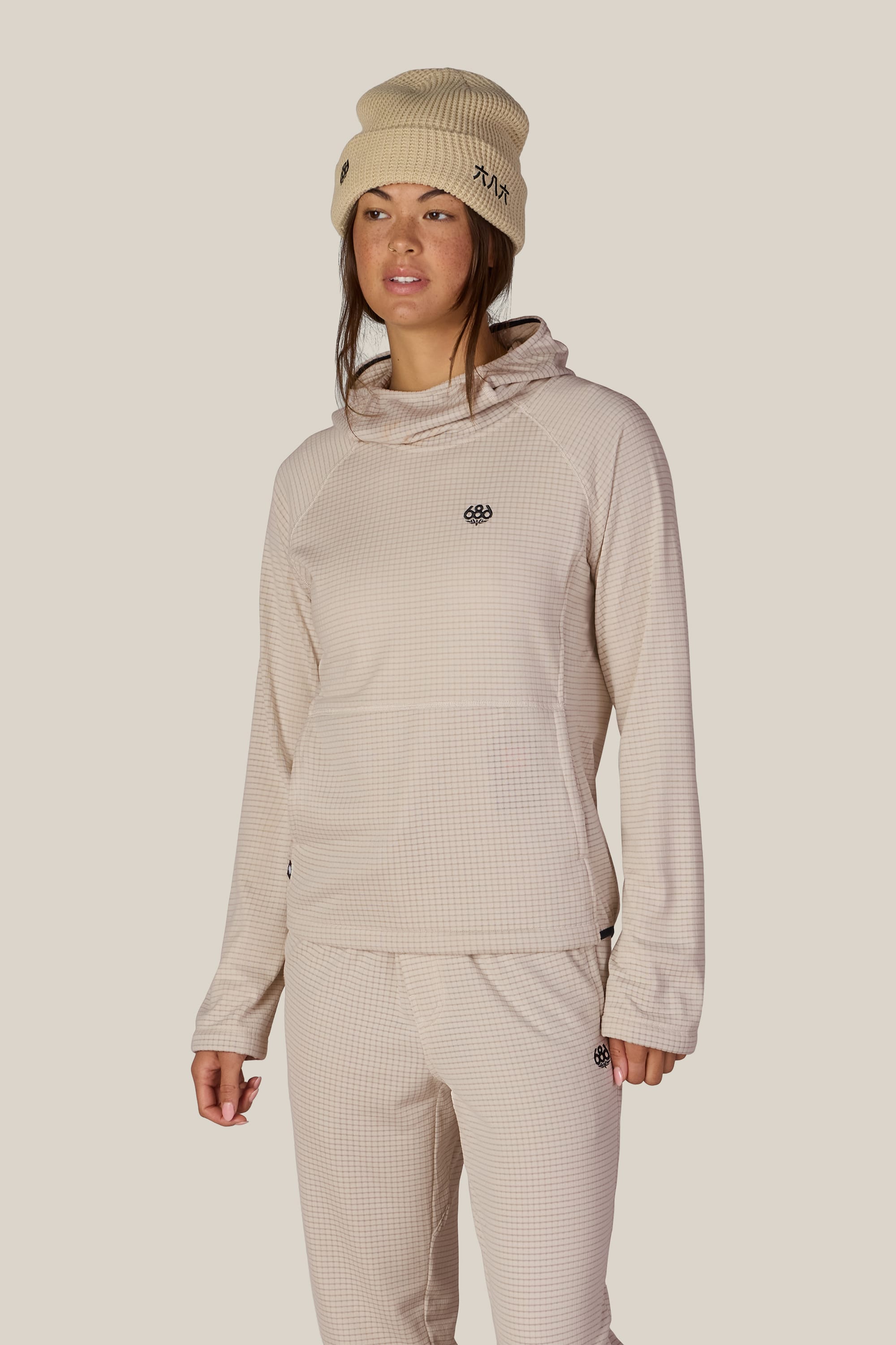 A woman wearing a beige beanie and pants, styled with the 686 Women’s Mission Grid™ Hoody by 686 and a women’s polyester jacket, stands against a plain light background.