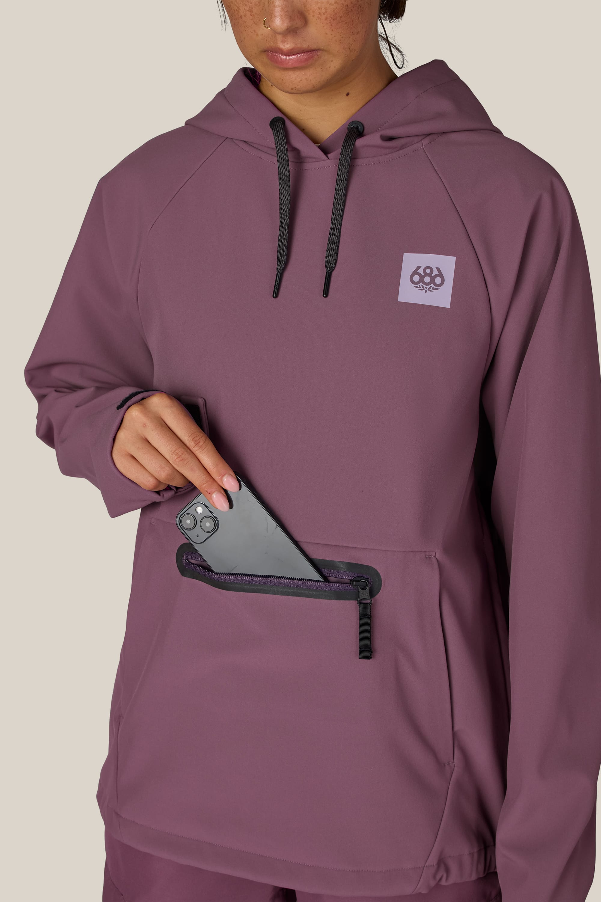 A woman wearing the 686 Women’s Waterproof Hoody places a smartphone into its zippered front pocket.
