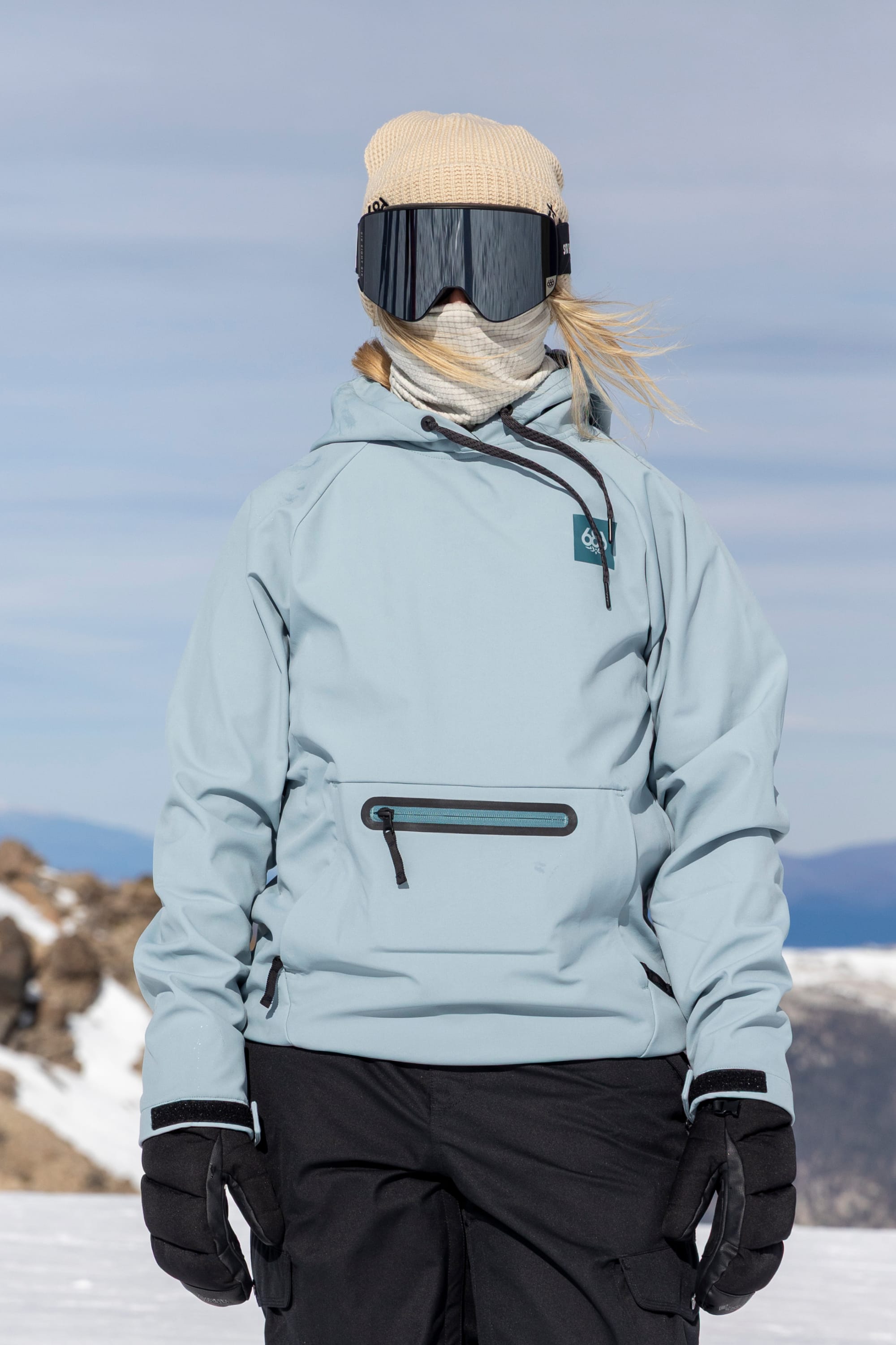 A person wearing the 686 Women’s Waterproof Hoody by 686, paired with black snow pants, gloves, a beige beanie, ski goggles, and a scarf stands outdoors in a snowy, mountainous landscape under a cloudy sky.