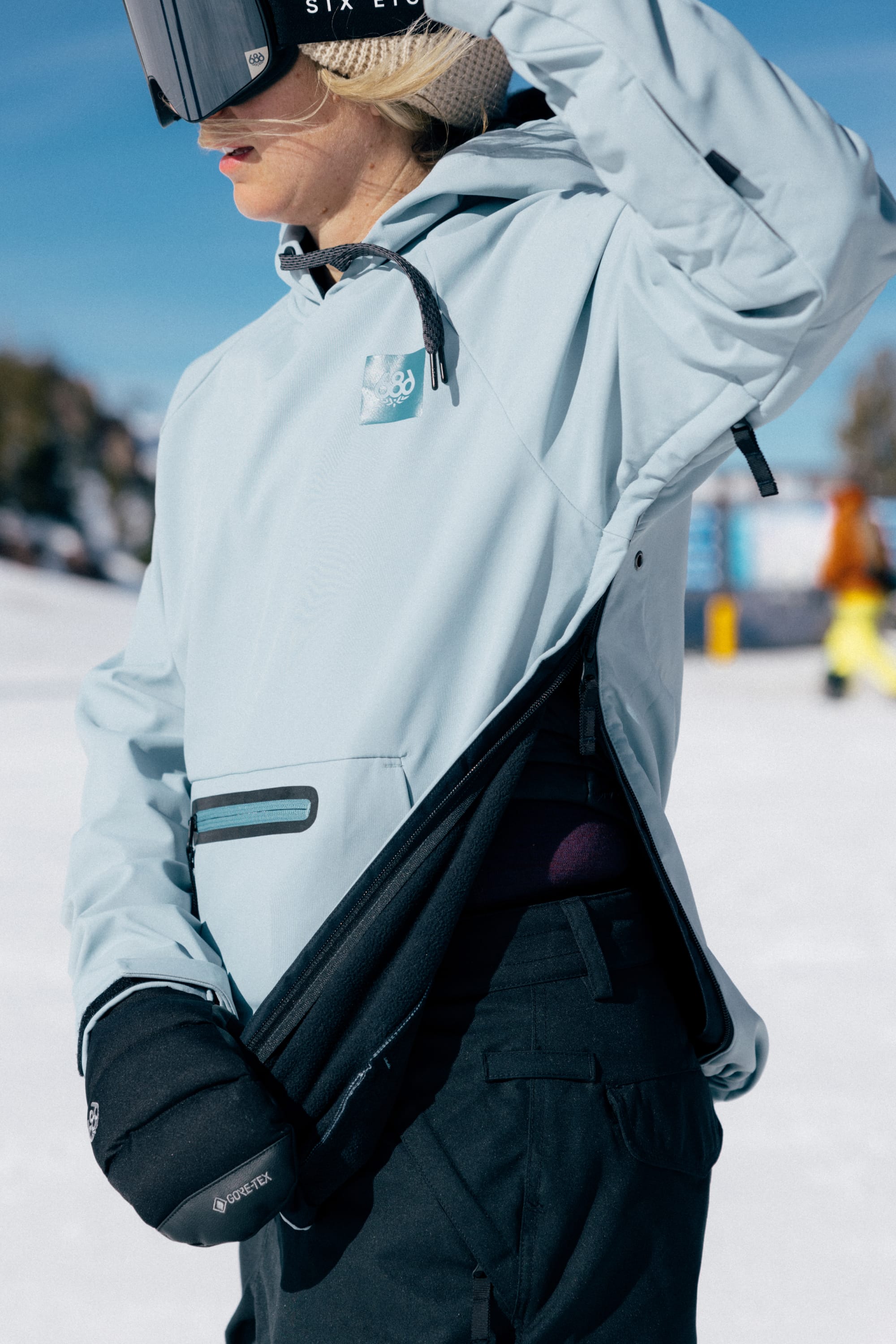 A person wearing the 686 Women’s Waterproof Hoody, along with black gloves and ski goggles, adjusts their jacket in a snowy outdoor setting on a sunny day.