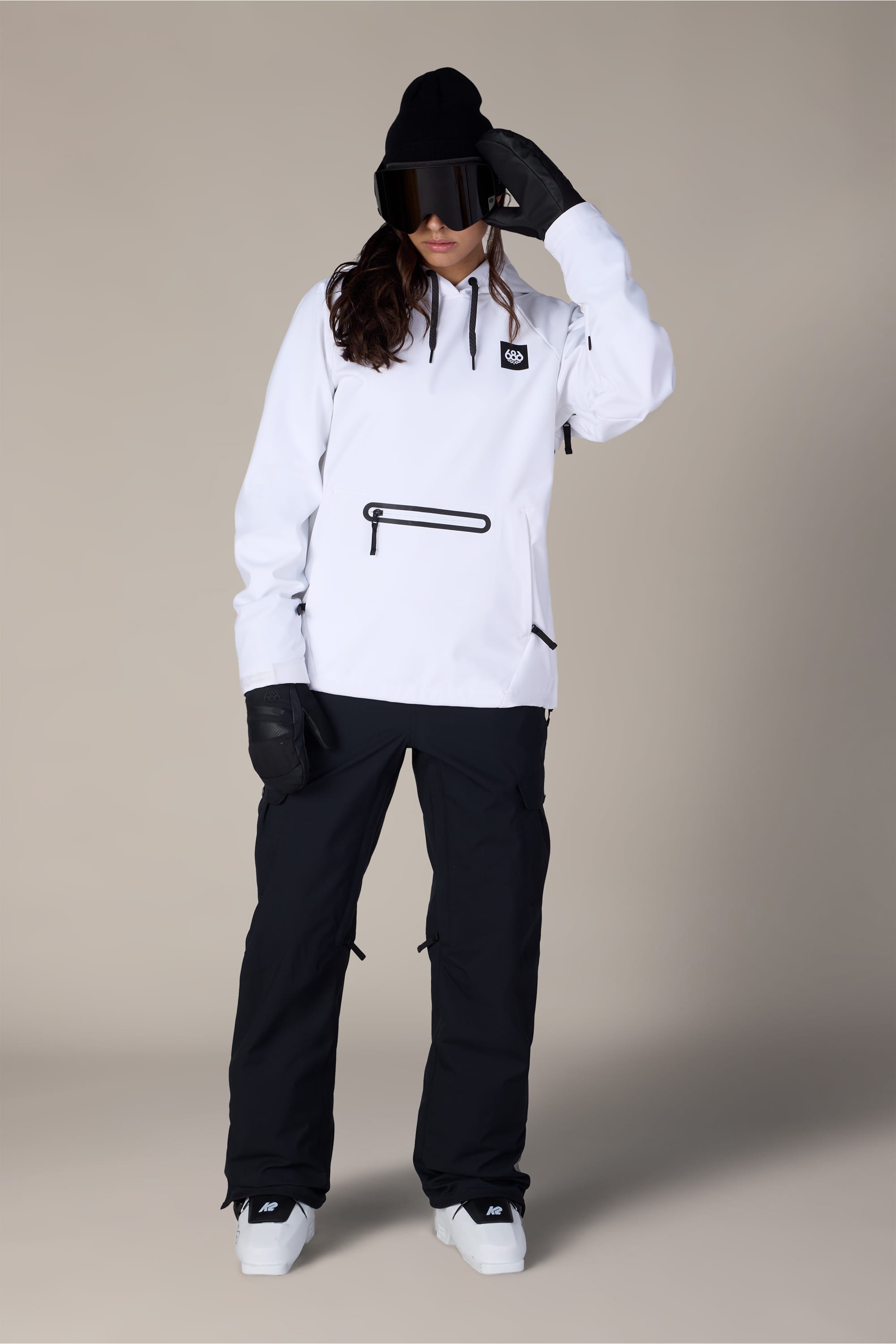 A person is wearing the 686 Women’s Waterproof Hoody by 686, paired with black snow pants, black gloves, a black beanie, and large ski goggles, standing against a plain background.