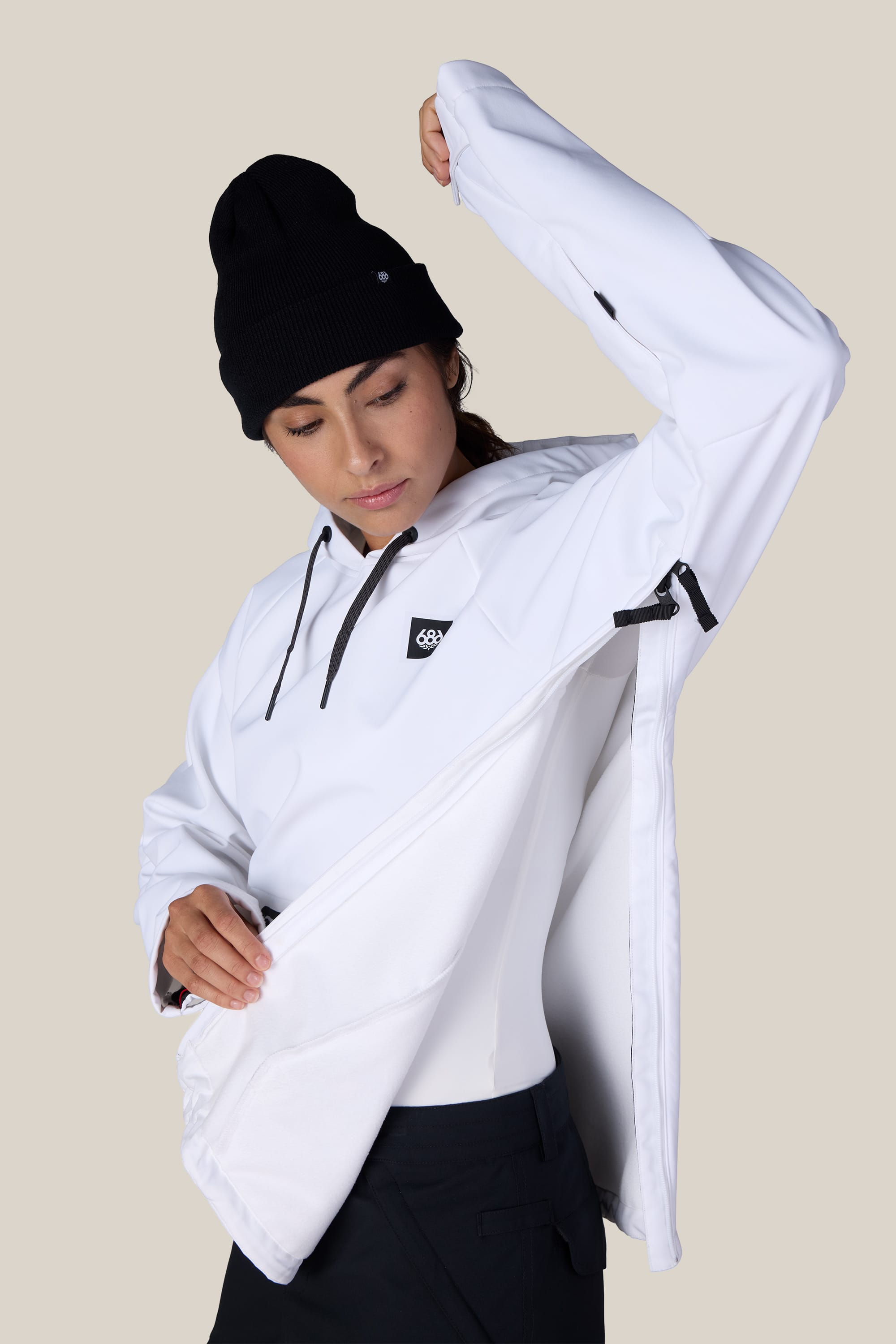 A woman wearing the 686 Women’s Waterproof Hoody in white and a black beanie opens a side zipper on the hoody to reveal its interior; she looks down slightly against a plain, light-colored background.