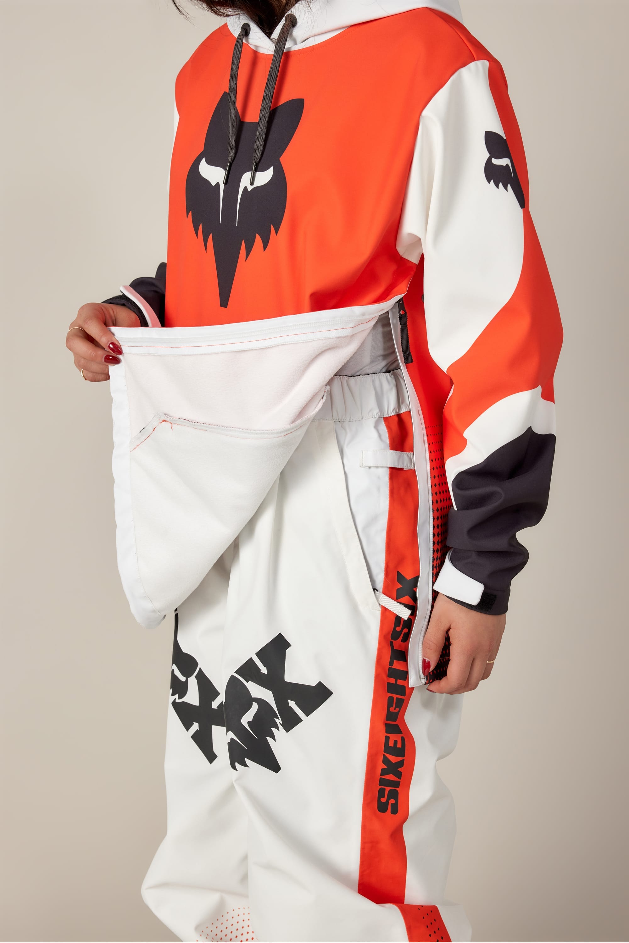 A person wearing the 686 Women's Fox Racing 97 Waterproof Hoody, featuring bold red, black, and white graphics and FOX branding, unzips a large front pocket on their pants with SKETCHY RY branding.