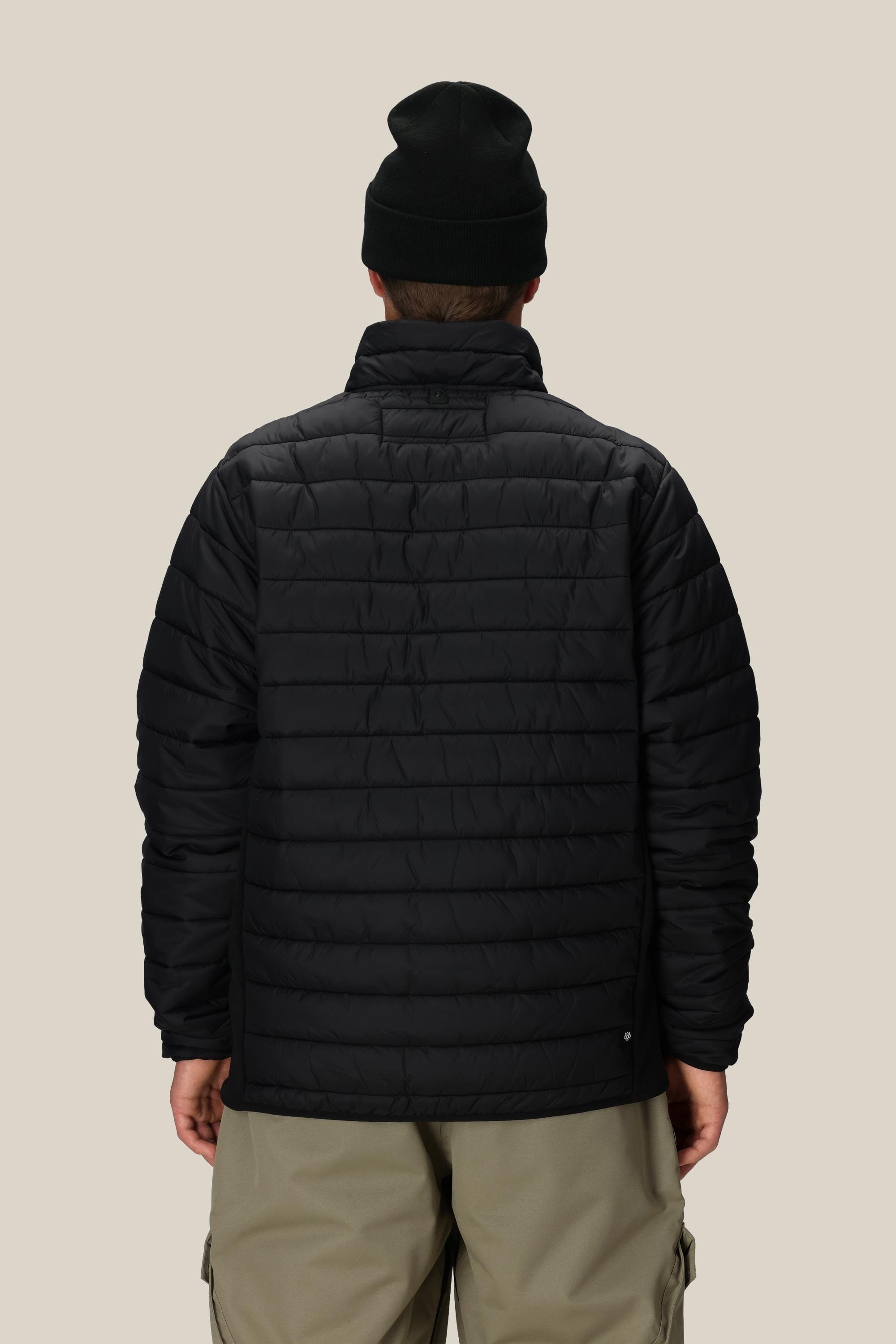 A person faces away from the camera wearing the 686 Mens Thermal Puff Jacket in black, a black beanie, and light-colored pants, standing against a plain beige background.