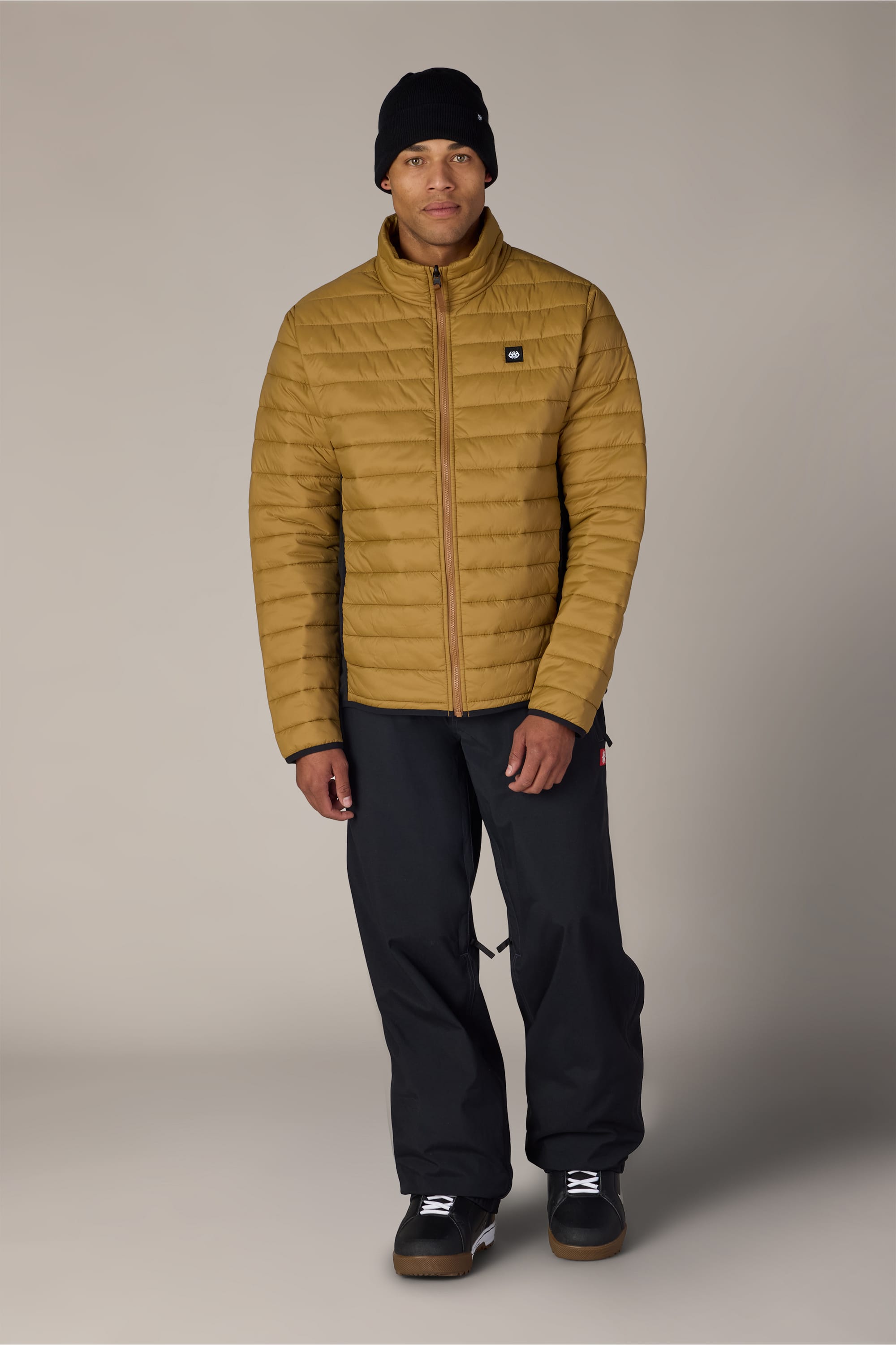 A person is wearing the 686 Men’s Thermal Puff Jacket in mustard yellow by 686, along with black pants, a black beanie, and black sneakers, standing facing forward against a plain, light background.