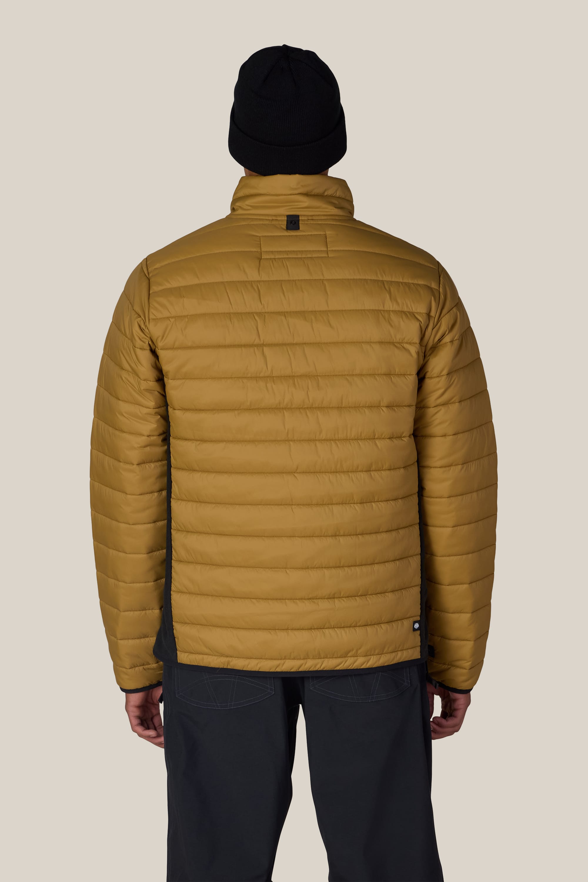 A person is shown from the back against a plain beige background, wearing the 686 Men’s Thermal Puff Jacket in mustard yellow by 686, paired with a black beanie and dark pants.