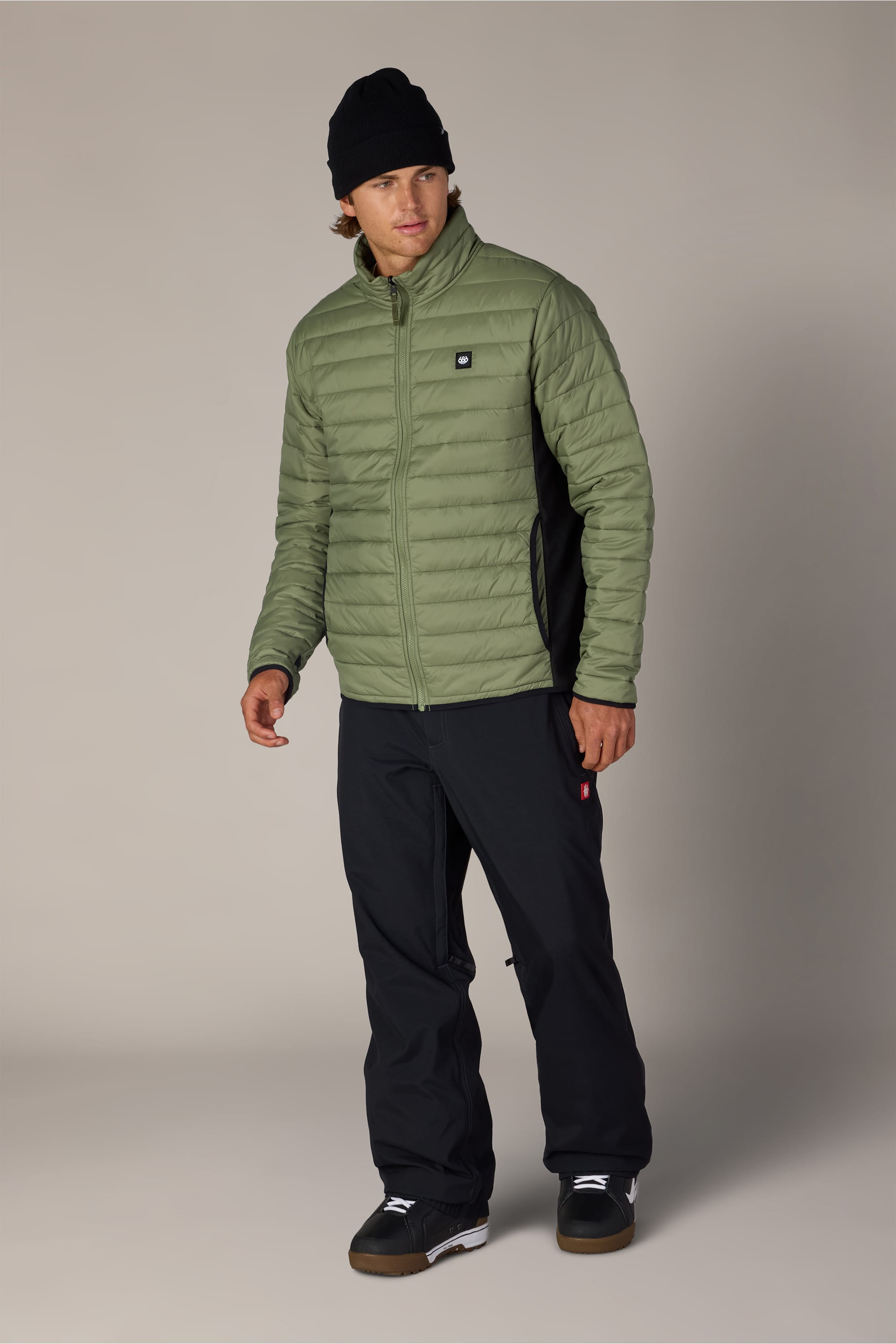 A person wearing the 686 Men’s Thermal Puff Jacket in green by 686, paired with black pants, a black beanie, and black shoes stands facing forward against a plain light background.