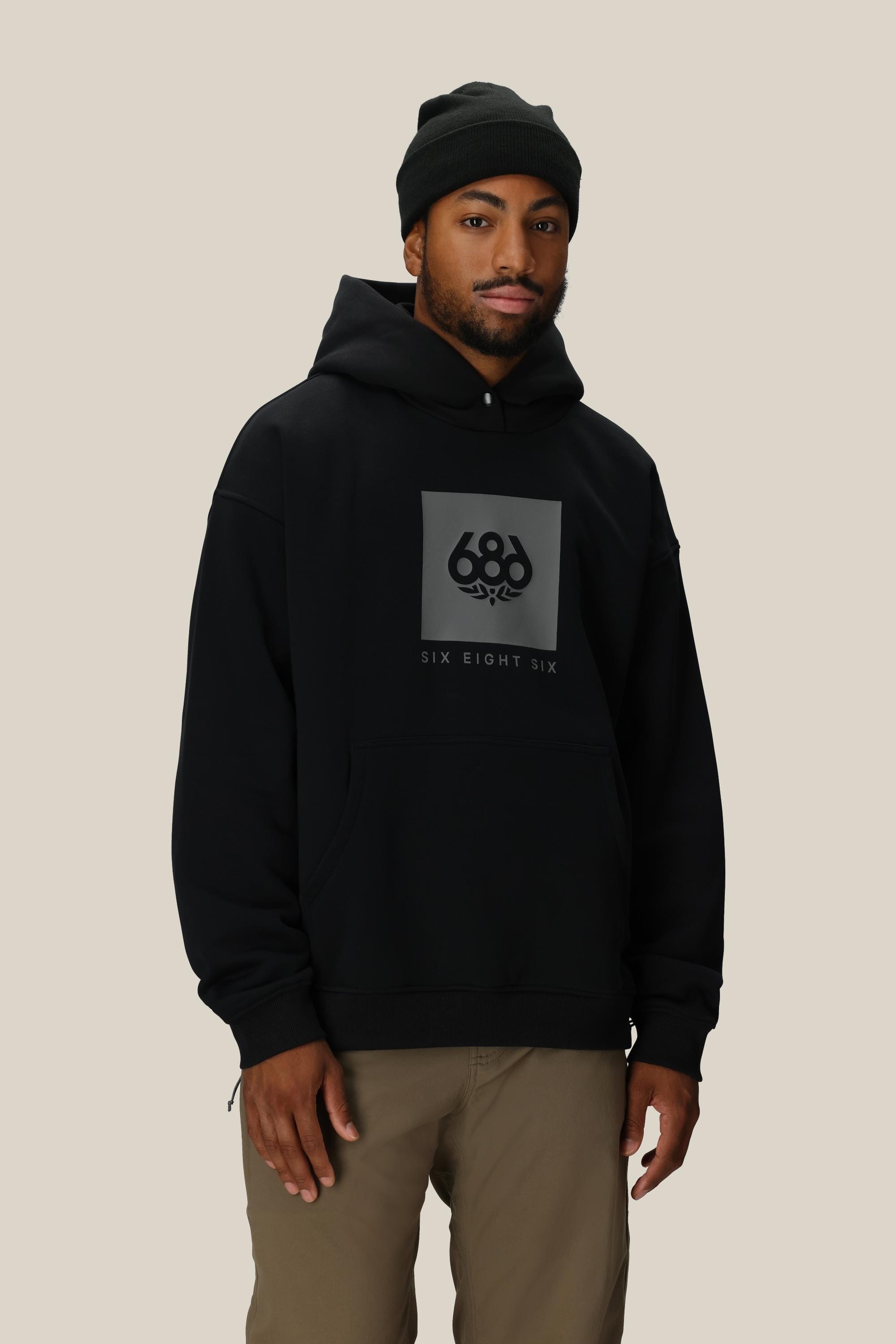 A man in a 686 Knockout Logo Premium Heavyweight Pullover Hoody and black beanie faces the camera with a neutral expression, wearing light brown pants against a beige background.