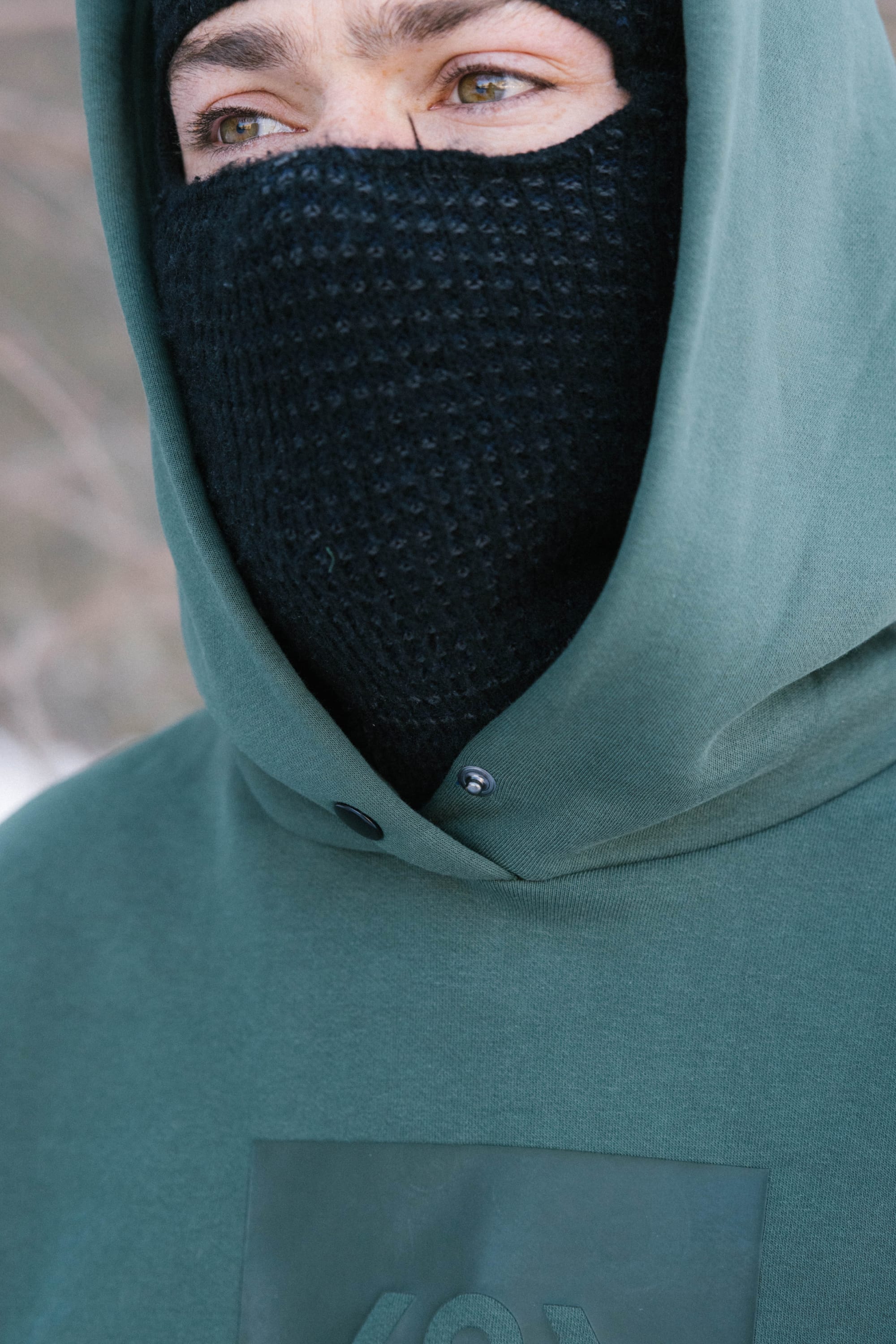 A person outdoors wears the 686 Knockout Logo Premium Heavyweight Pullover Hoody in green and a black knit face mask, with only their eyes and part of their nose visible against a blurred, likely cold background.