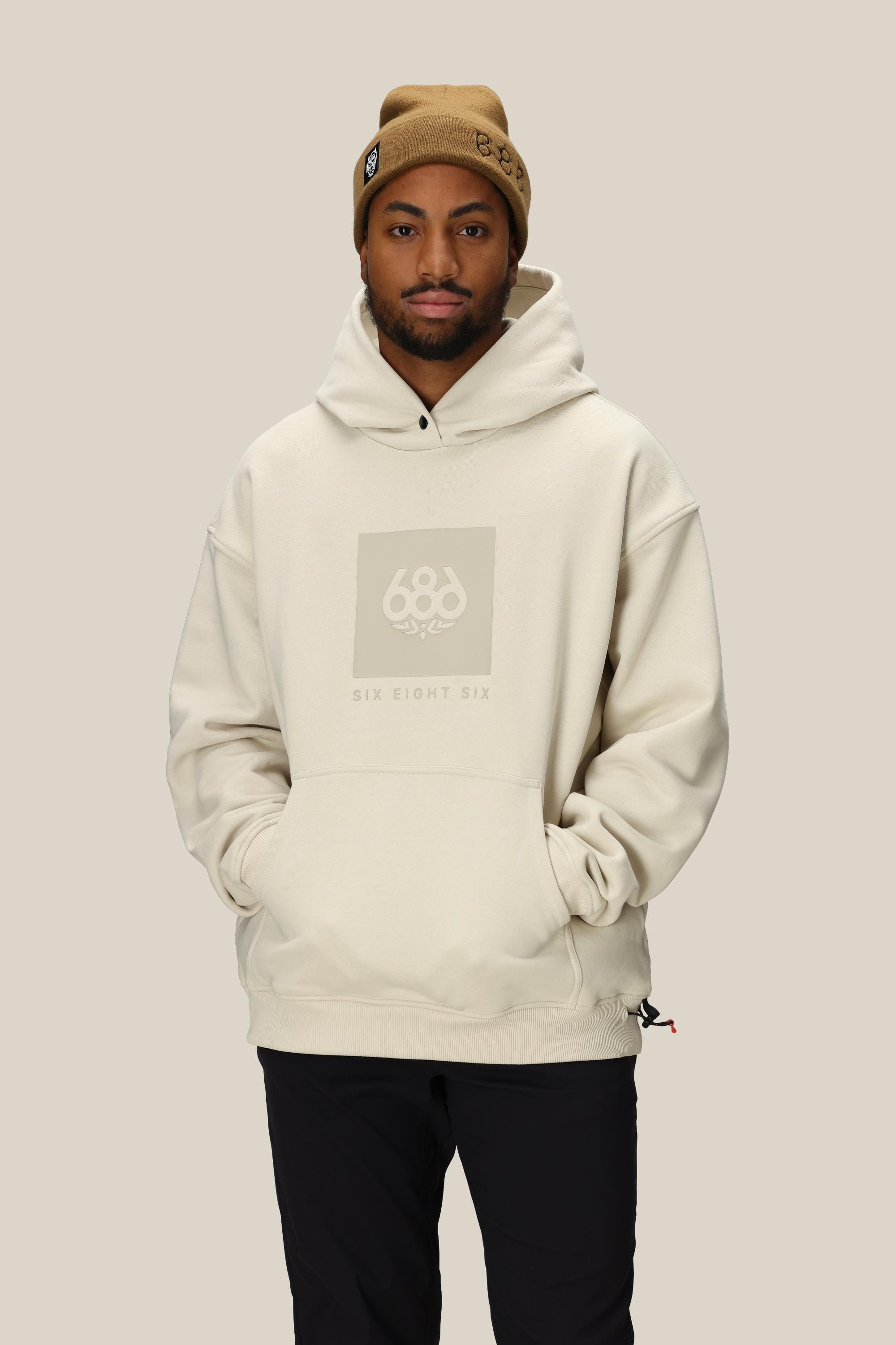 A man in a tan beanie and an oversized 686 Knockout Logo Premium Heavyweight Pullover Hoody stands against a plain beige background with his hands in the hoodie pocket.