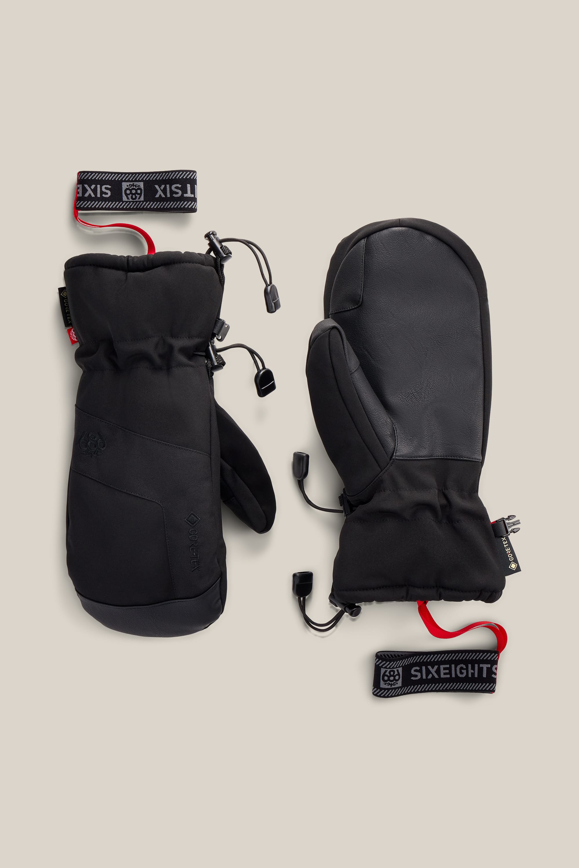 A pair of black 686 Men’s GORE-TEX® Linear™ Mitts with reinforced palms and adjustable wrist straps, featuring white SIXEIGHT branding on black; the mittens include red tags and drawstrings for adjustment, and are shown against a plain beige background.