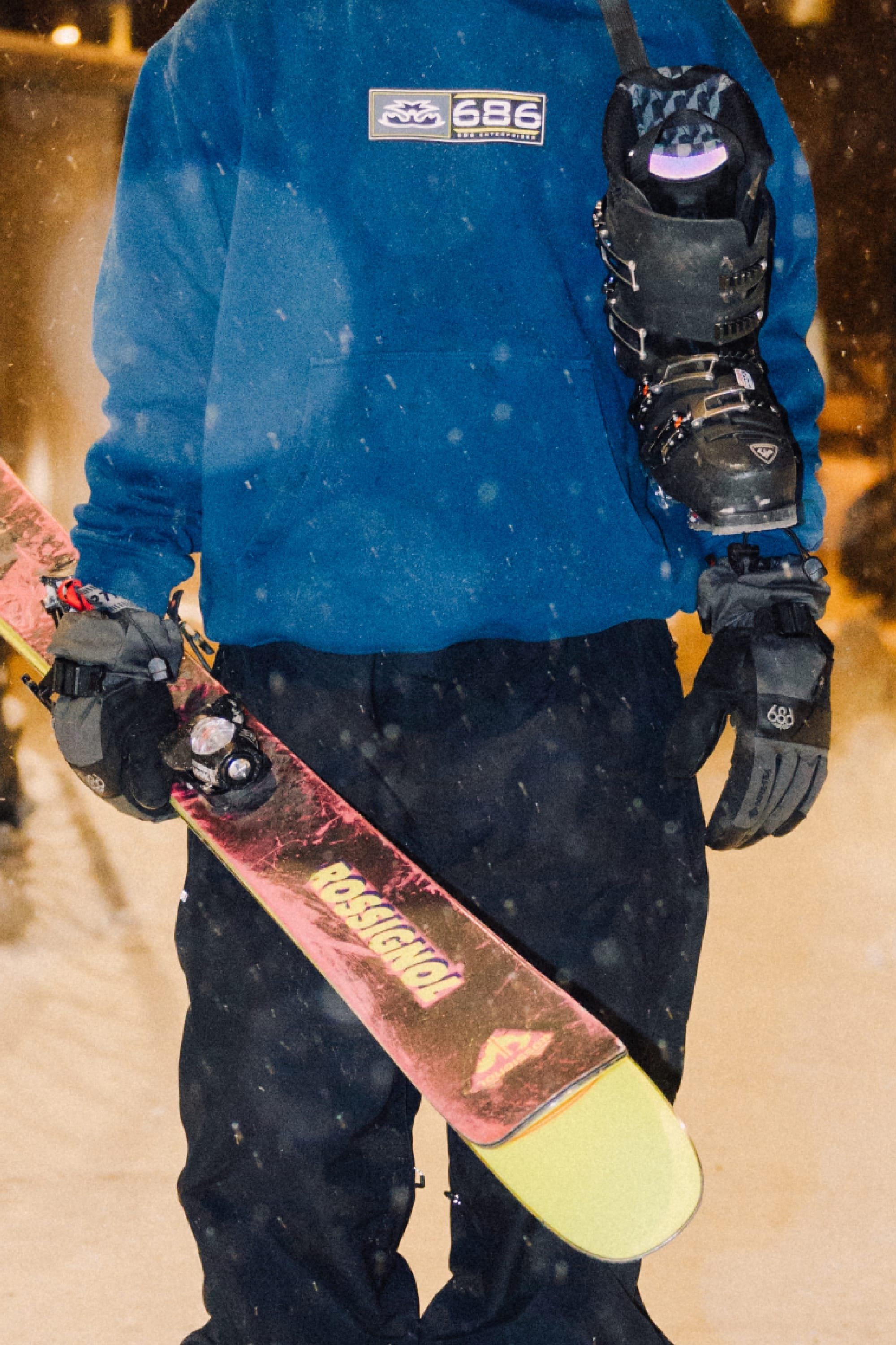 A man in a blue sweatshirt and black pants holds red and yellow skis. Hes wearing 686 Mens GORE-TEX Smarty 3-In-1 Gauntlet Gloves by 686 and has a black helmet slung over his shoulder. Snow falls softly in the background.