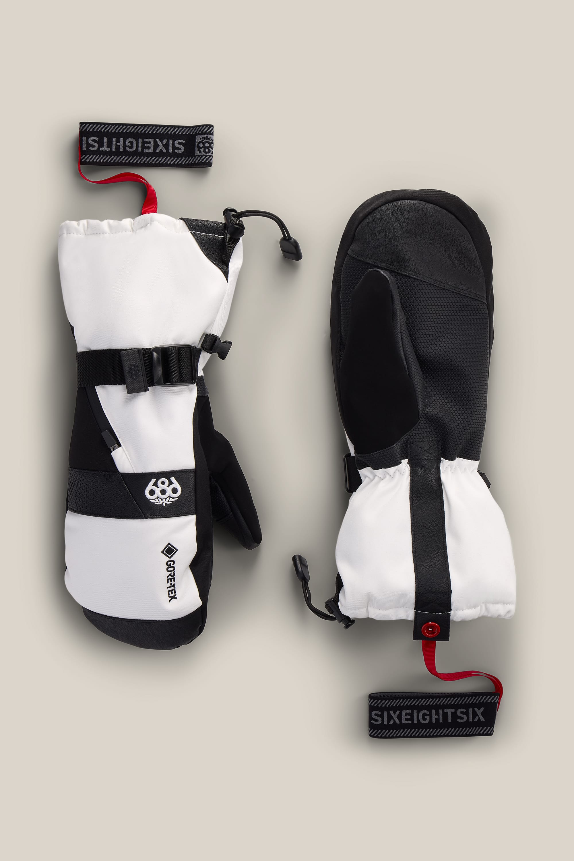 A pair of 686 Men’s GORE-TEX® SMARTY® 3-In-1 Gauntlet Mitts in black and white, with black palms, adjustable straps, red SIXEIGHTSiX wrist leashes, and prominent 686 and GORE-TEX logos, displayed on a beige background.
