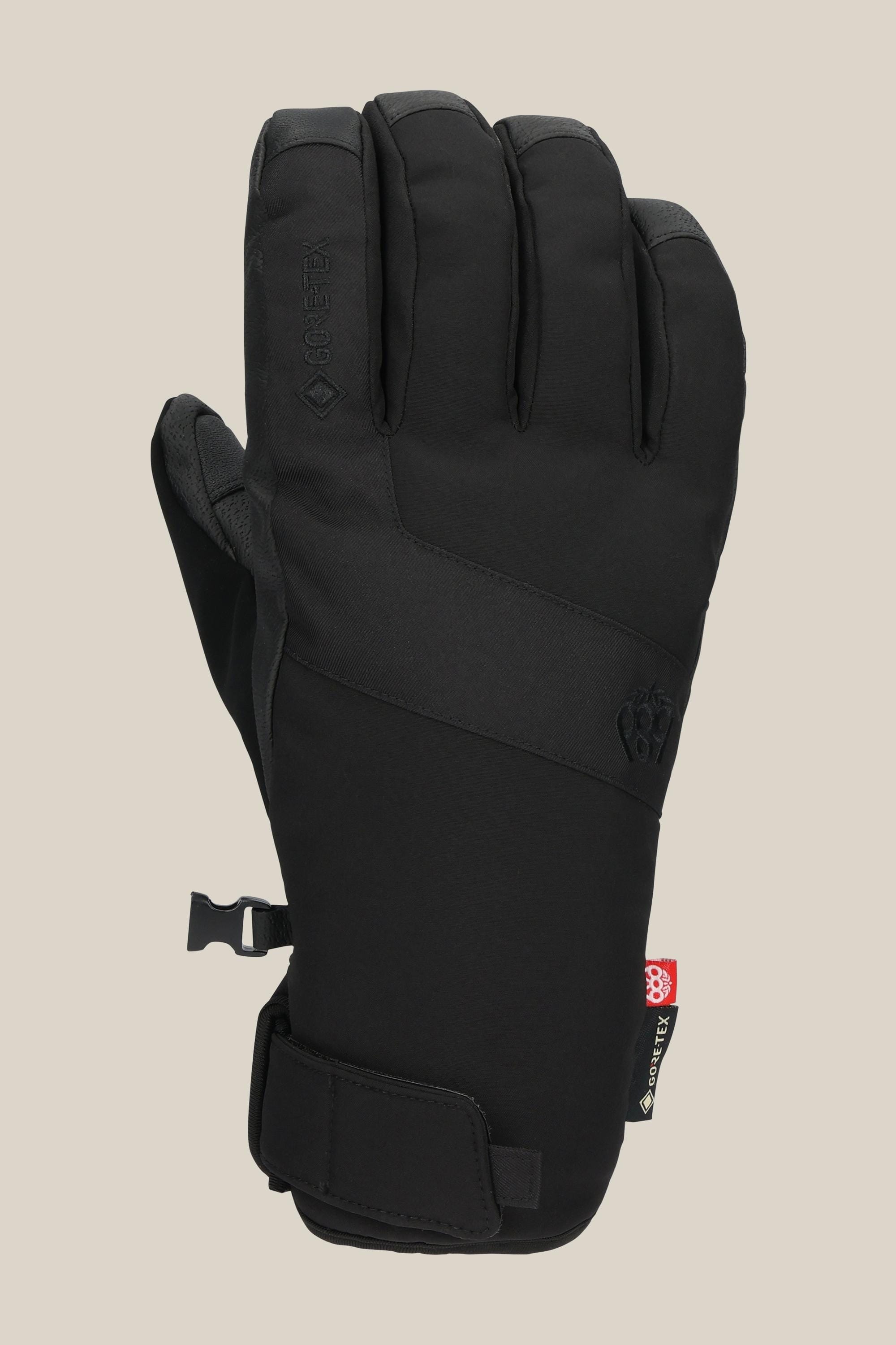 The 686 Mens GORE-TEX Linear Under Cuff Glove in black features a Velcro wrist strap, subtle 686 branding on the back, and a plastic side clip—this sleek PU glove is shown against a beige background.
