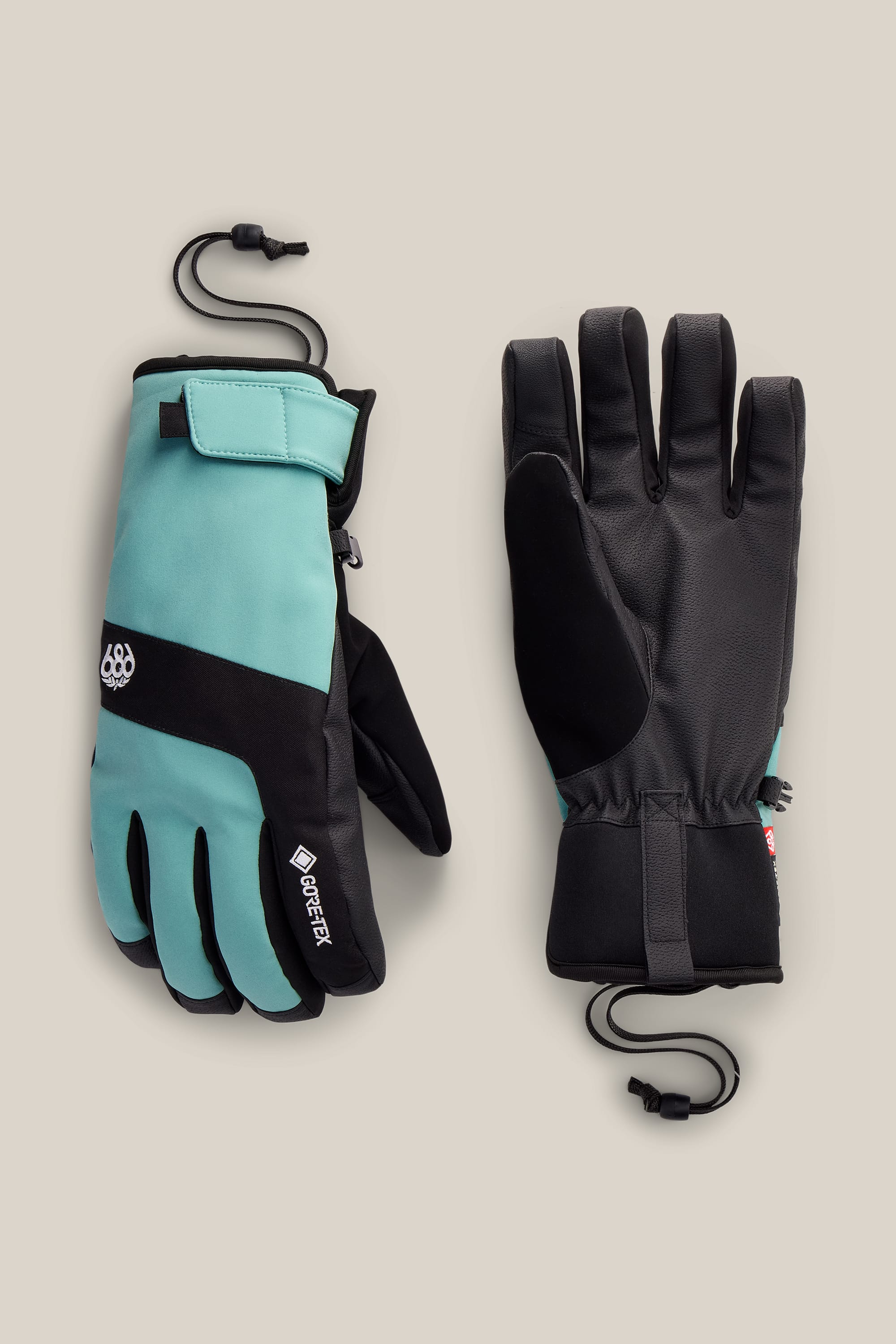 A pair of 686 Men’s GORE-TEX® Linear™ Under Cuff Gloves in teal and black, featuring adjustable wrist straps and cinch cords, are displayed on a beige background. One glove reveals the palm, while the other shows the branded back.