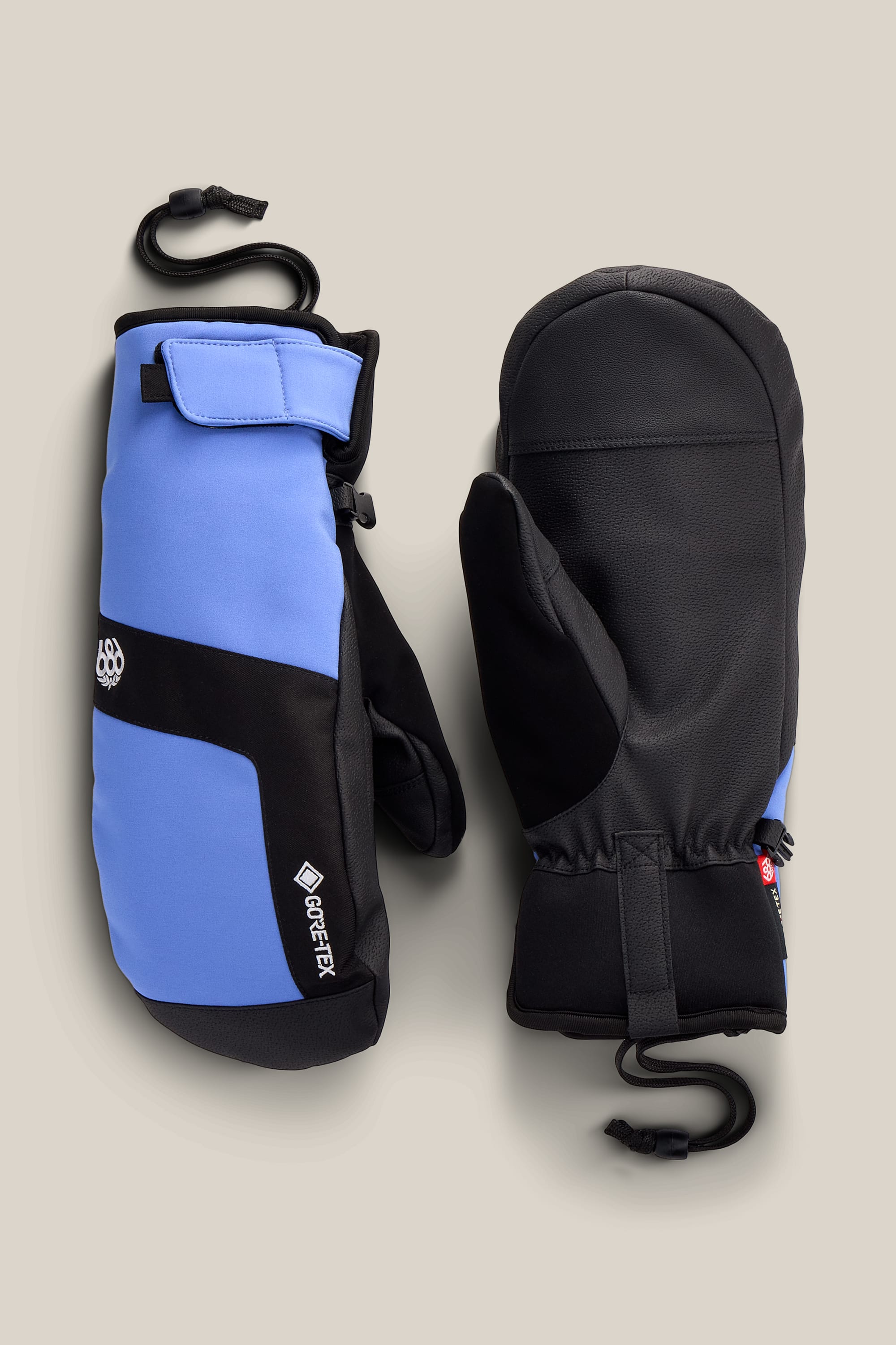 A pair of 686 Men’s GORE-TEX® Linear™ Under Cuff Mitts in blue and black, featuring wrist straps, are displayed on a light beige background with one mitten showing the palm side and the other showing the back side.