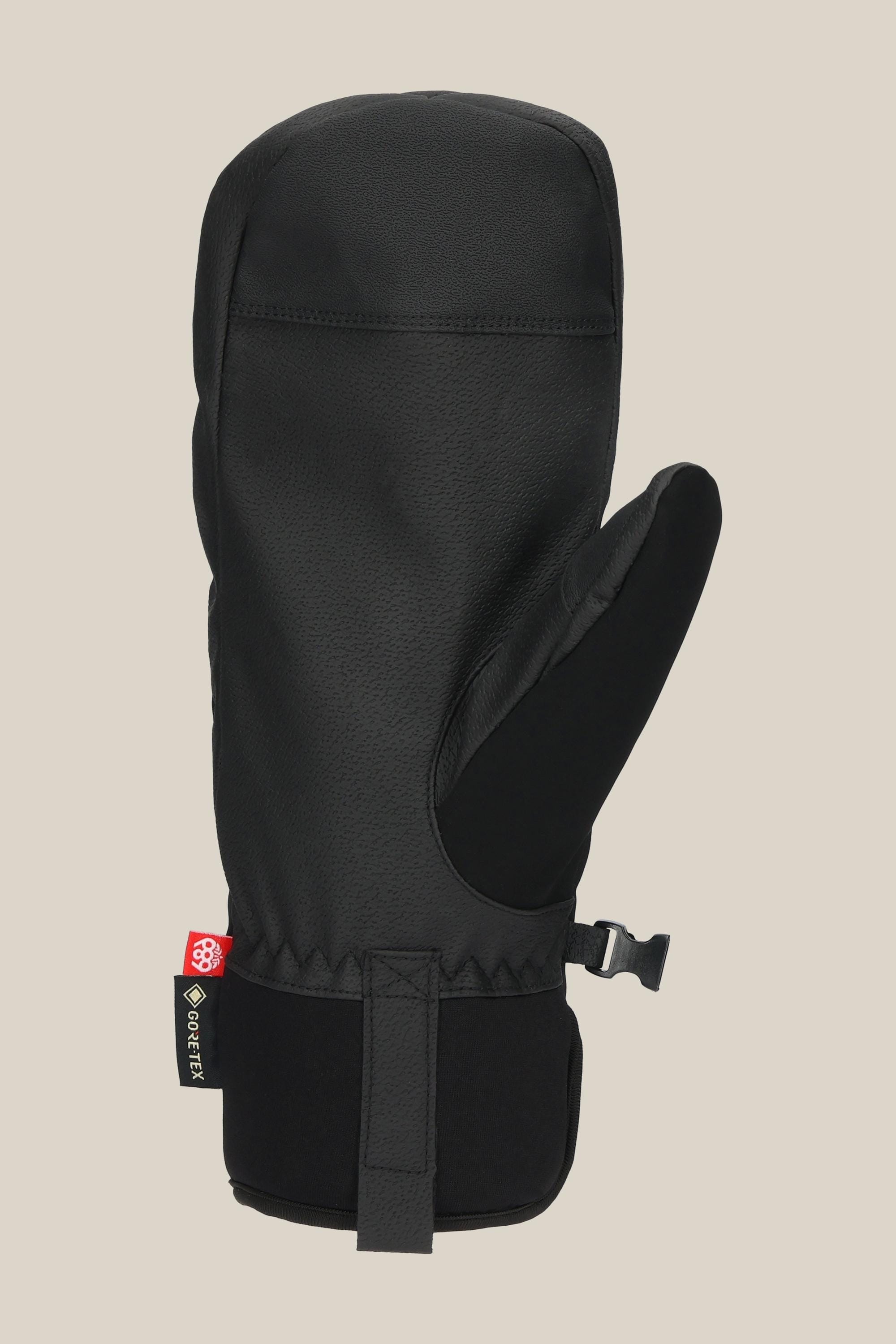 The 686 Mens GORE-TEX Linear Under Cuff Mitt, in black with a textured finish, features an adjustable wrist strap and a small red-and-white logo tag on the cuff, shown upright against a plain beige background.