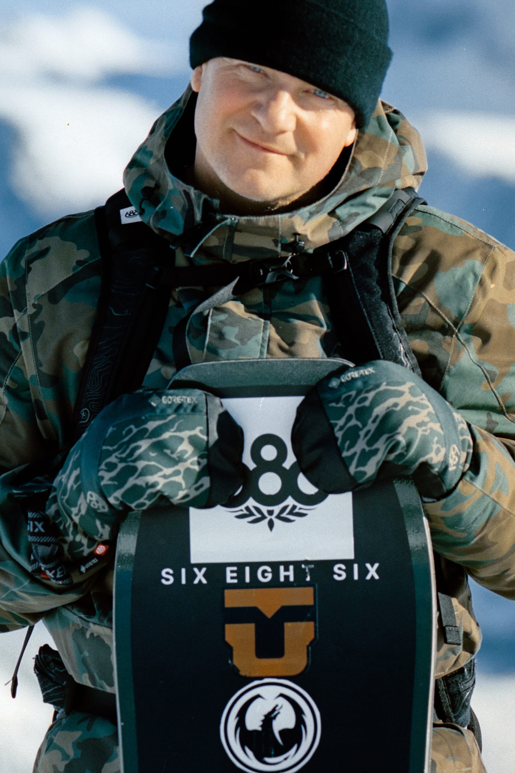 A person in a black beanie, camouflage jacket, and 686 Mens GORE-TEX Linear Under Cuff Mitt holds a vertically positioned snowboard with SIX EIGHT SIX logos, set against snowy terrain in the background.