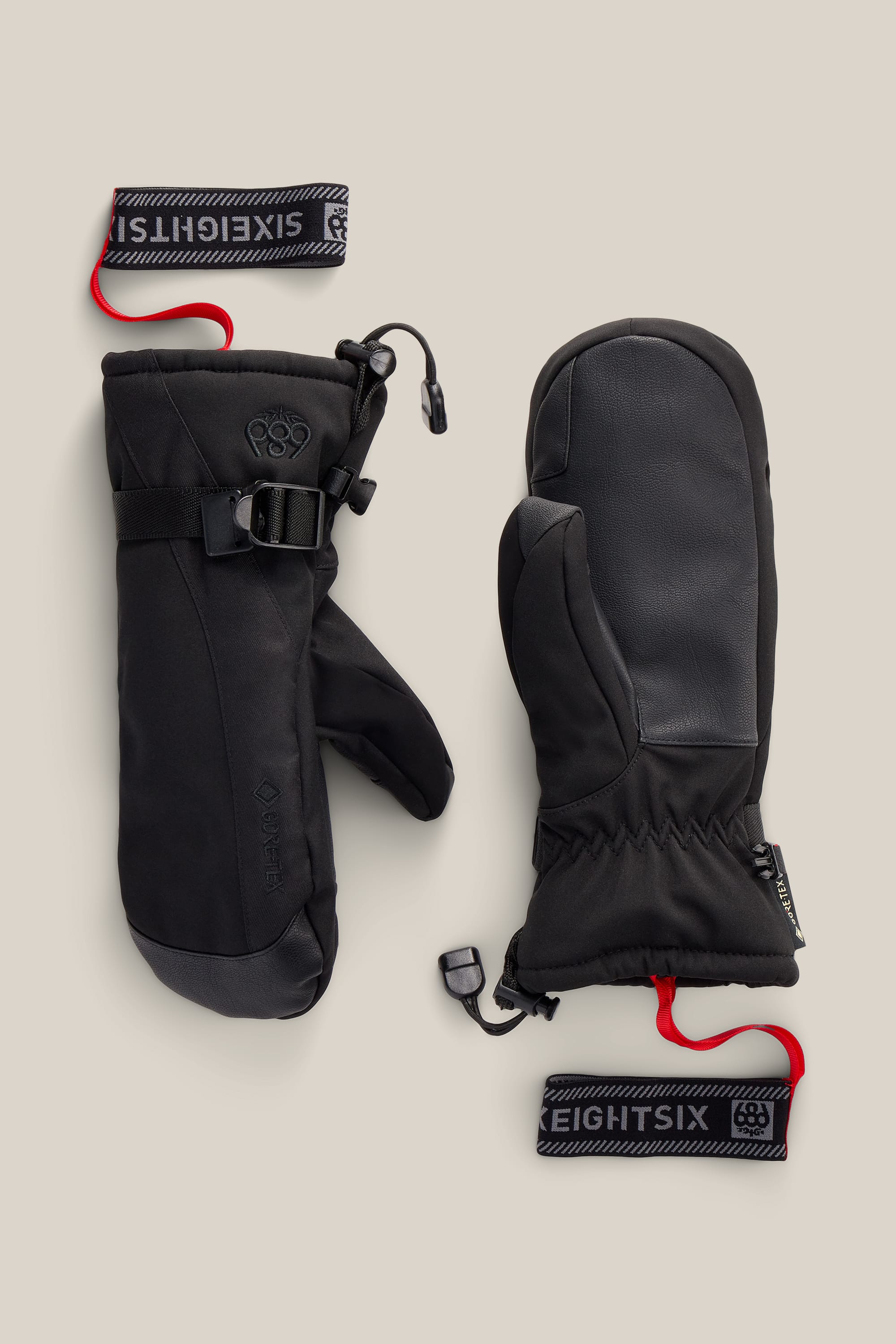 A pair of 686 Women’s GORE-TEX® Linear™ Mitts in black, featuring insulated construction with wrist straps, drawcords, leather palms, and red leash attachments, displayed flat on a neutral background.