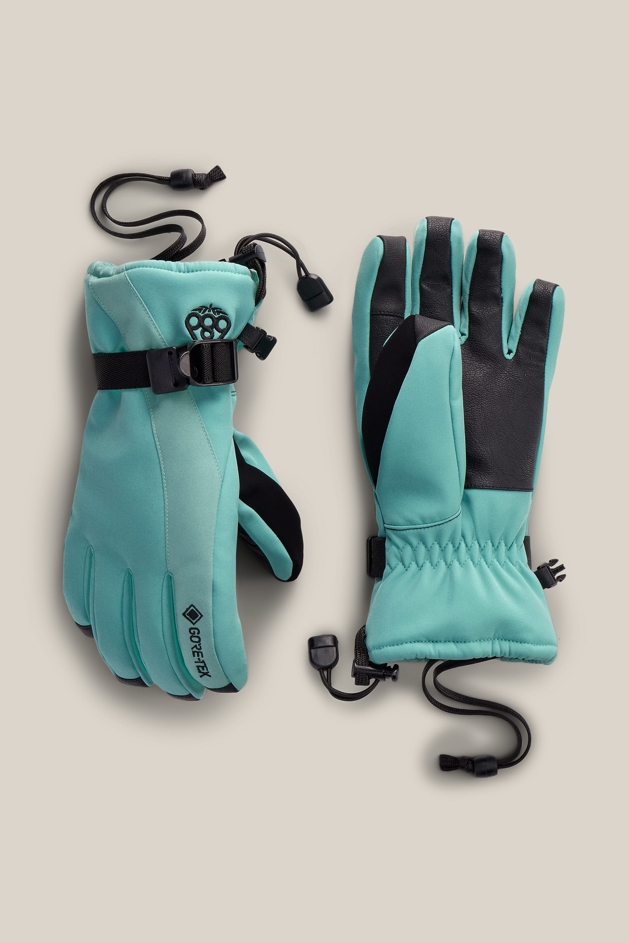 A pair of 686 Women’s GORE-TEX® Linear™ Gloves in teal, featuring black adjustable straps, black palms, wrist leashes, and a visible GORE-TEX label, are shown laid flat on a beige background.