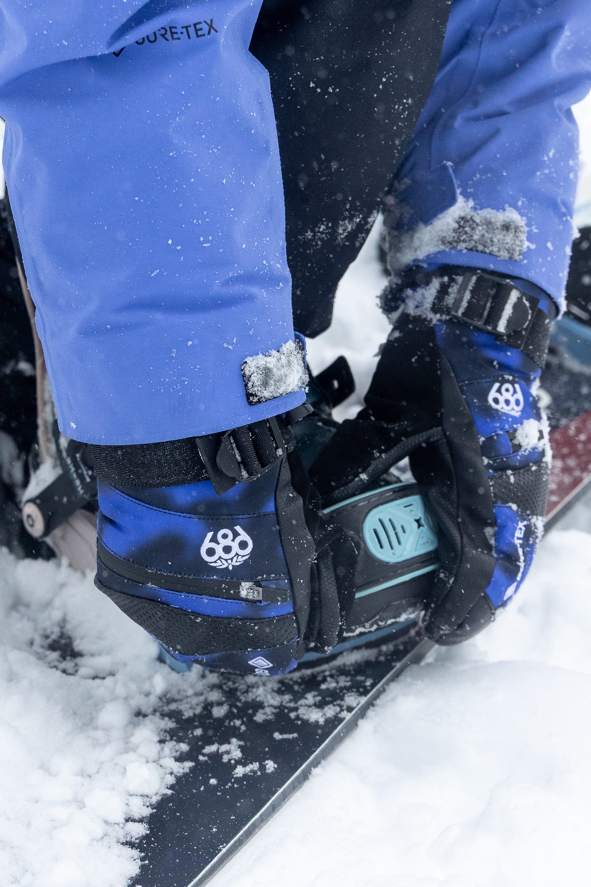 A person wearing the 686 Women’s GORE-TEX® SMARTY® 3-In-1 Gauntlet Mitt fastens a snowboard binding in the snow.