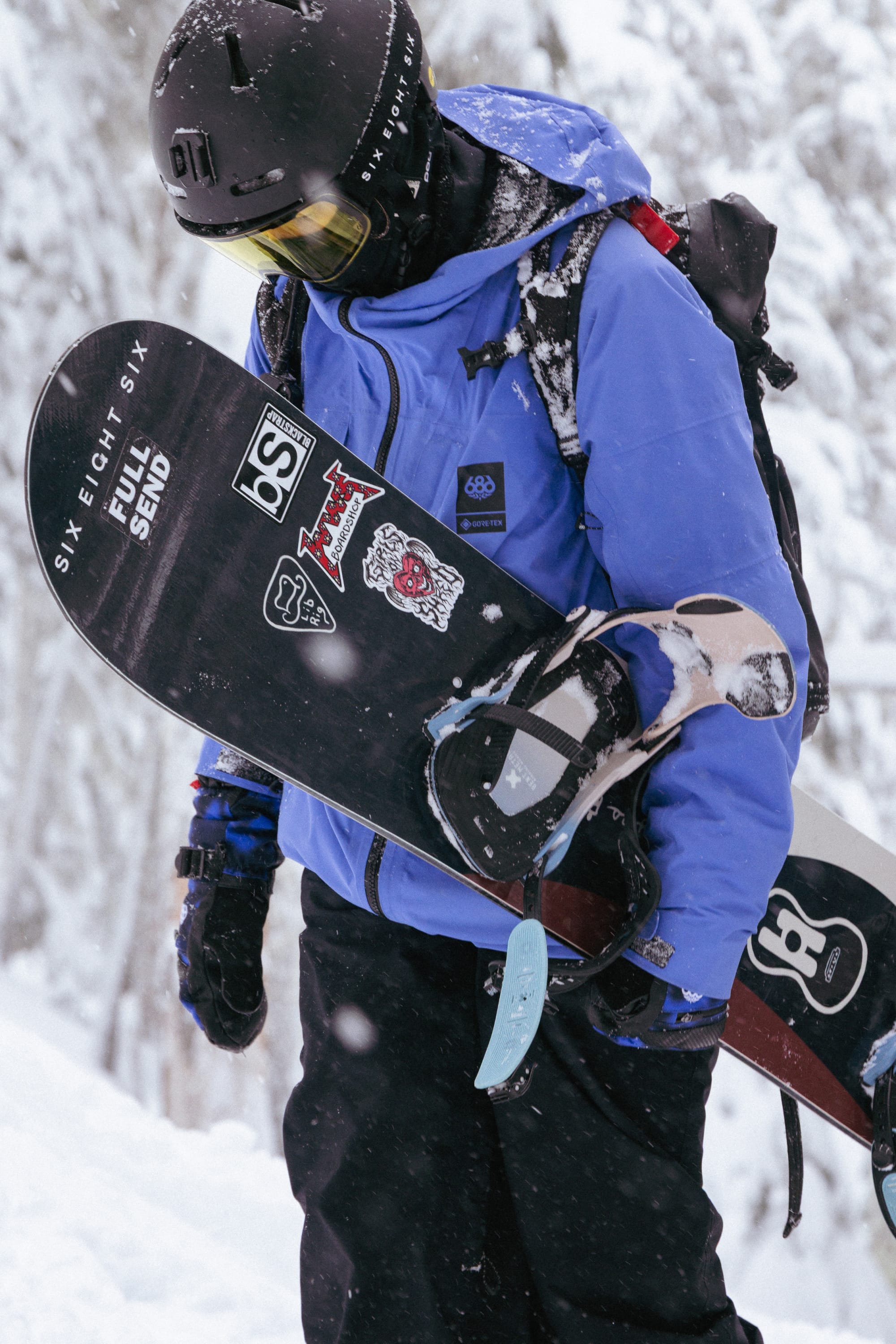 Snowboarder in a blue jacket and helmet, wearing 686 Women’s GORE-TEX® SMARTY® 3-In-1 Gauntlet Mitts, holds a snowboard decorated with stickers while standing in a snowy forest with snowflakes falling and snow-covered trees.