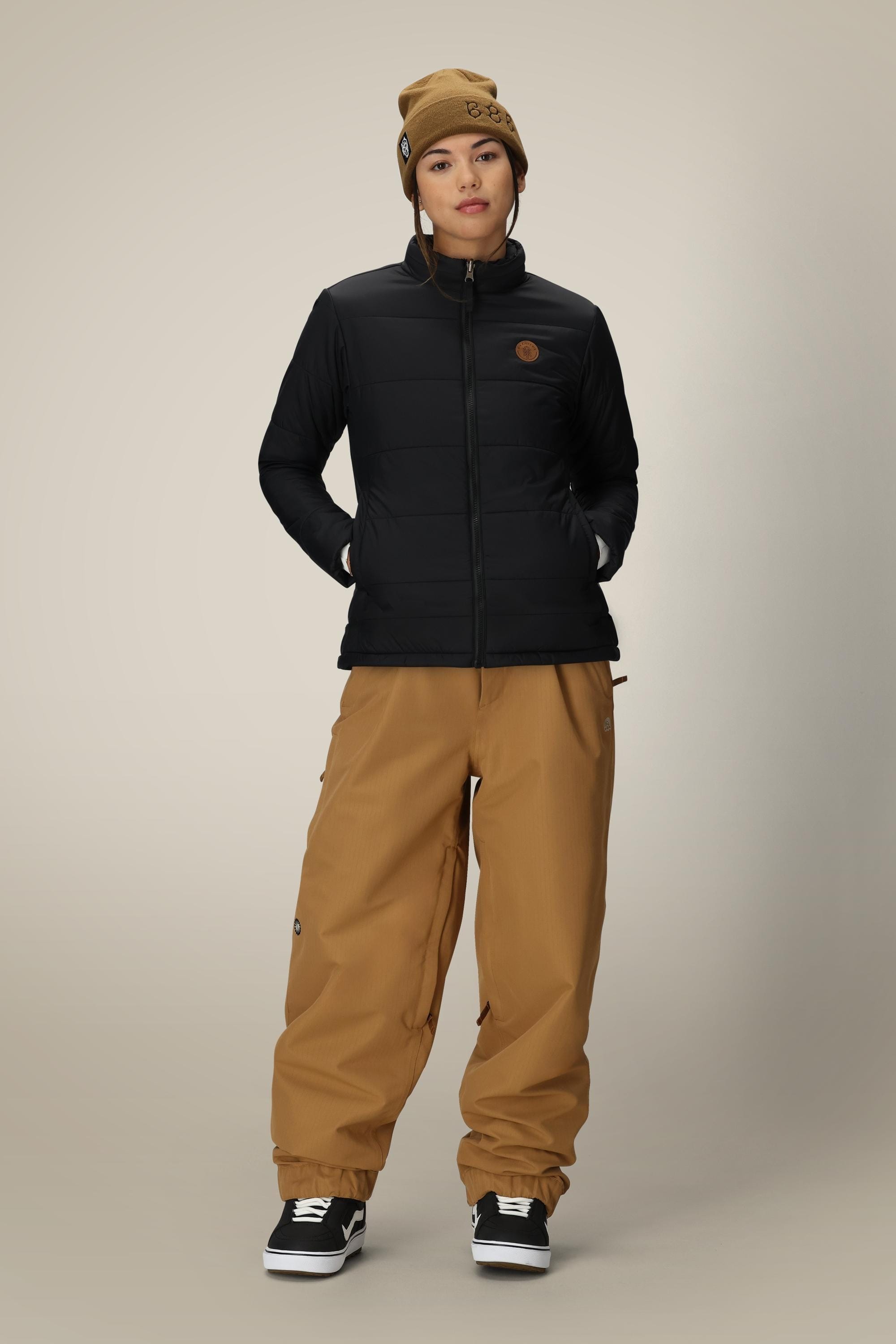 Wearing the 686 Women’s Thermal Puff Insulated Jacket by 686, a person stands with hands in pockets against a plain background, paired with a brown beanie, brown baggy pants, and black sneakers.