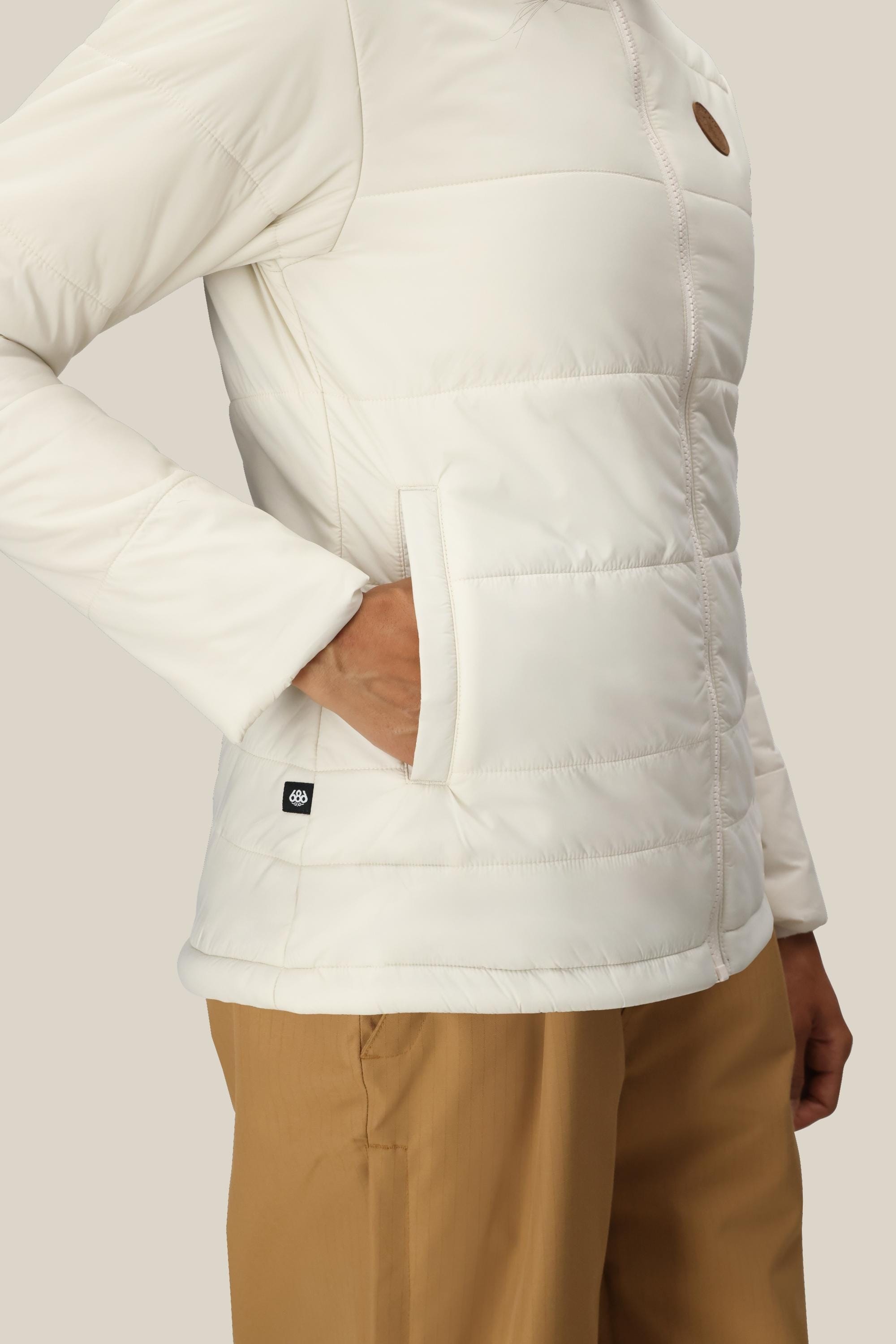 A woman wearing the 686 Women’s Thermal Puff Insulated Jacket with one hand in the pocket and tan pants stands against a neutral background.