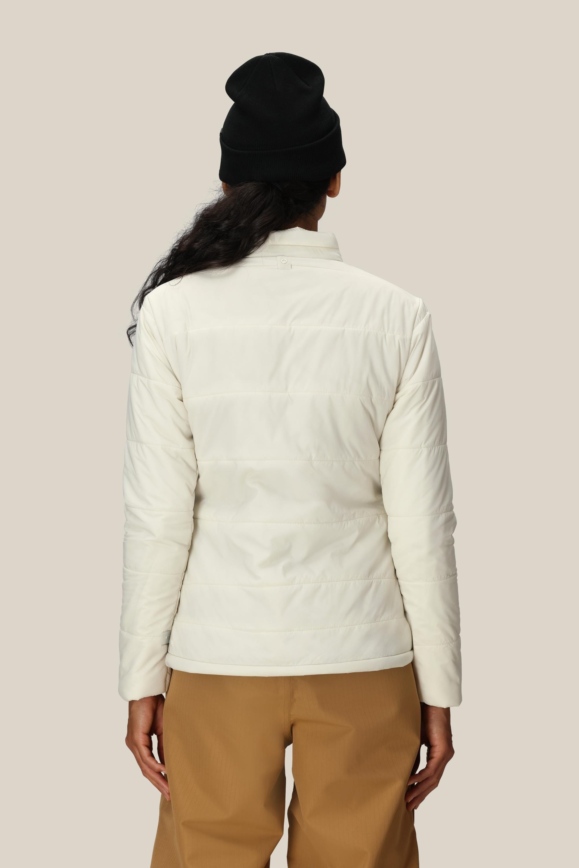 A person is shown from the back, standing against a plain beige background and wearing a black beanie, tan pants, and the 686 Women’s Thermal Puff Insulated Jacket by 686 in white.