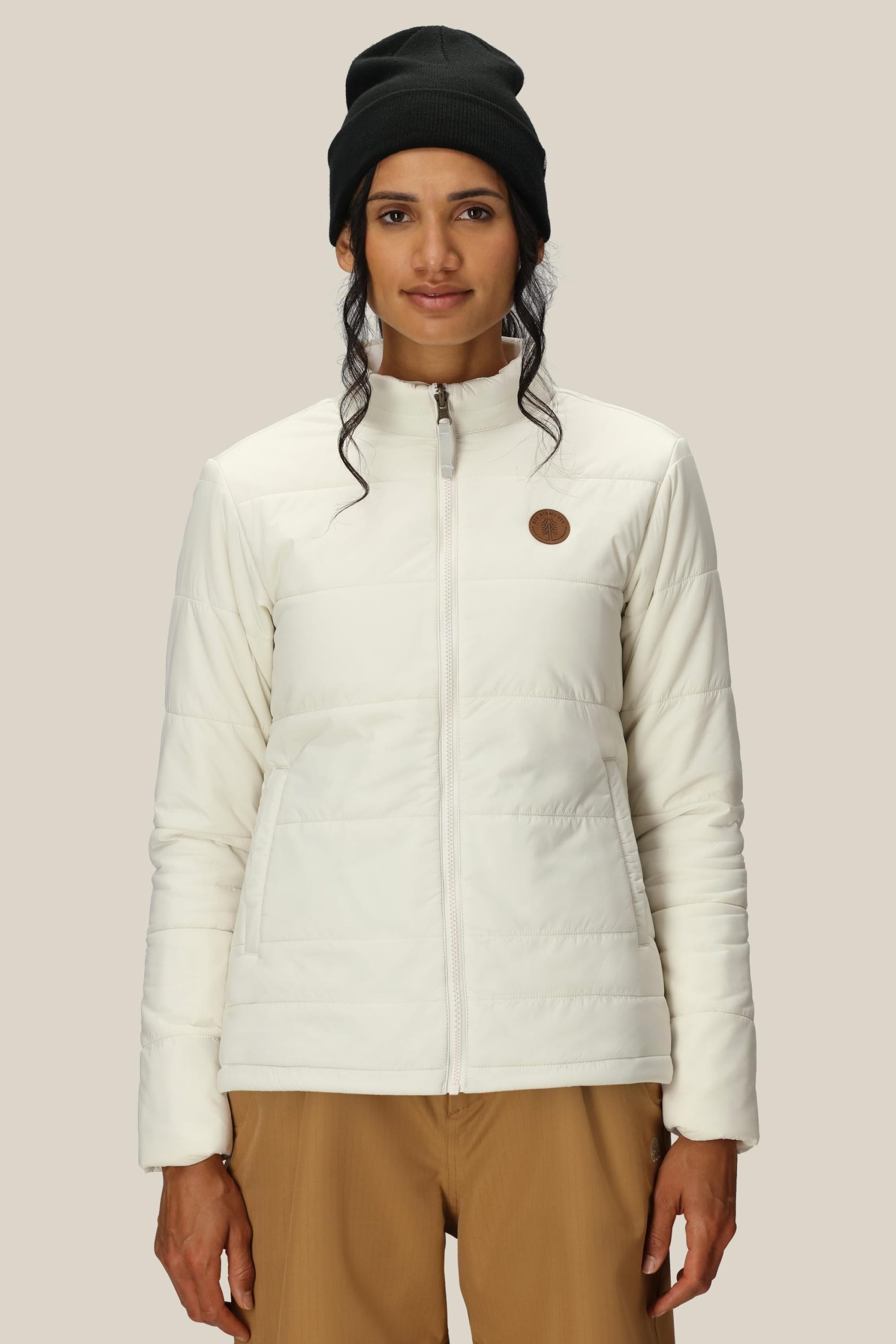 A person faces forward and smiles slightly, wearing a black beanie, light brown pants, and the 686 Womens Thermal Puff Insulated Jacket—a white polyester jacket with a round logo on the chest—against a plain light background.