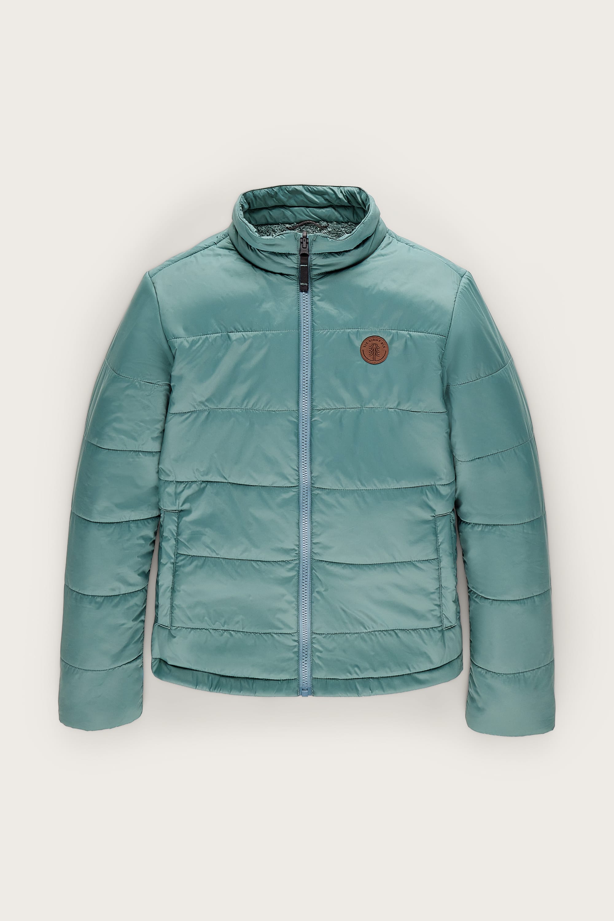 The 686 Women’s Thermal Puff Insulated Jacket by 686 is a teal puffer featuring a high collar, front zipper, side pockets, and a round brown patch on the left chest, displayed against a plain light background.