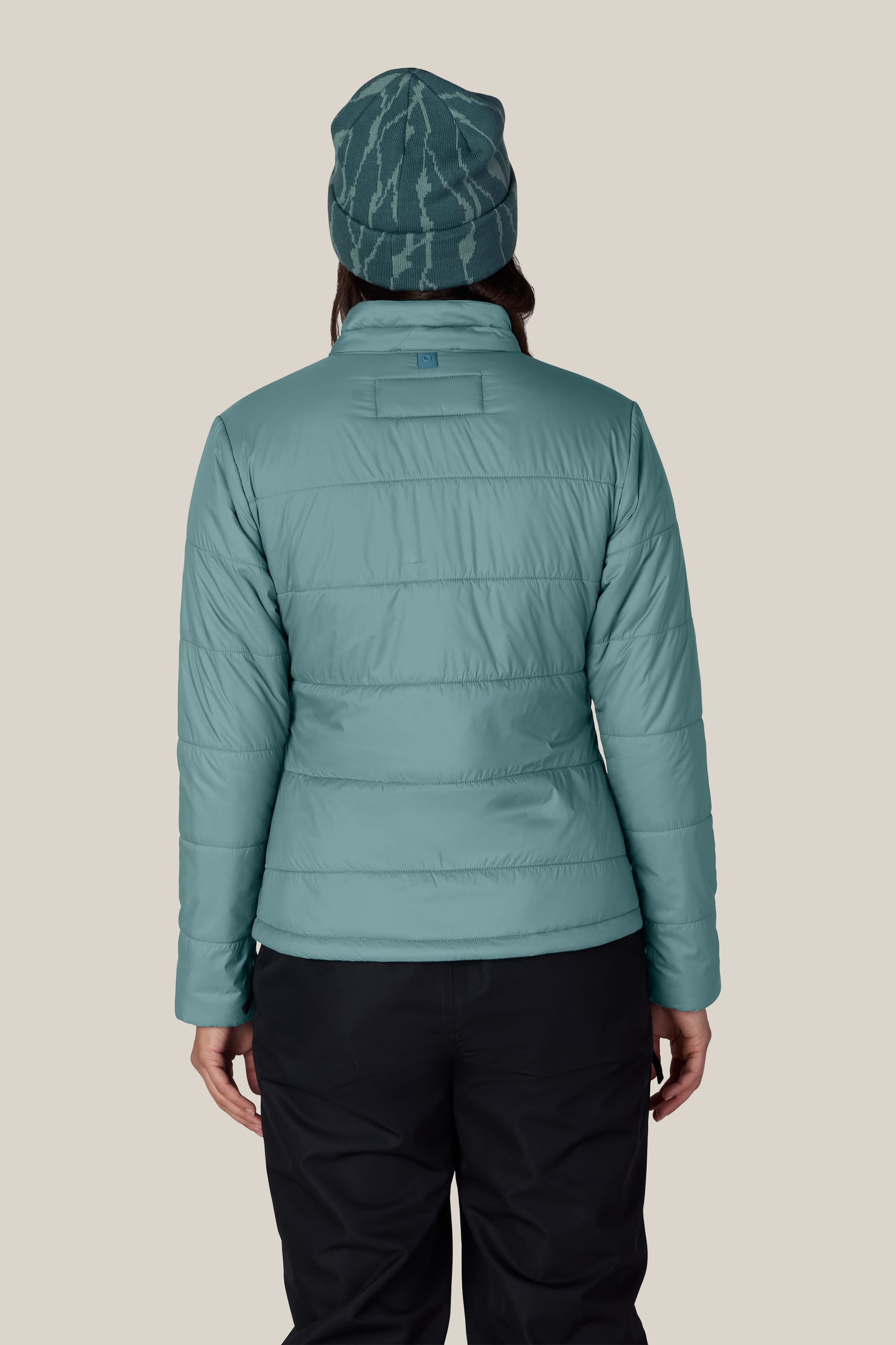 A person is shown from the back against a plain beige background, wearing the 686 Women’s Thermal Puff Insulated Jacket in teal and a patterned teal beanie.
