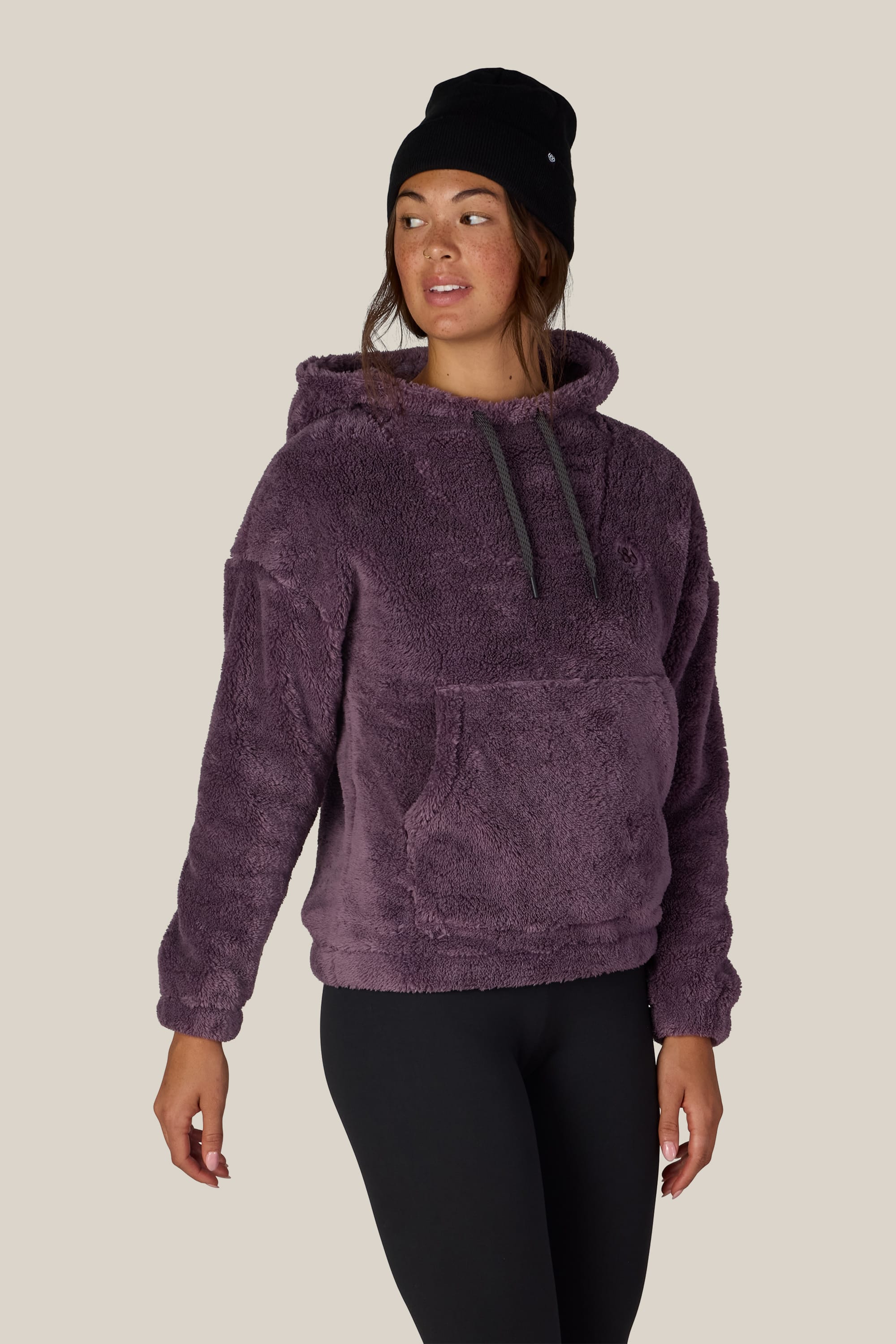 A person wearing the 686 Women’s Sherpa Hoody in purple by 686, paired with a black knit beanie and black leggings, stands against a plain beige background, looking to the side.