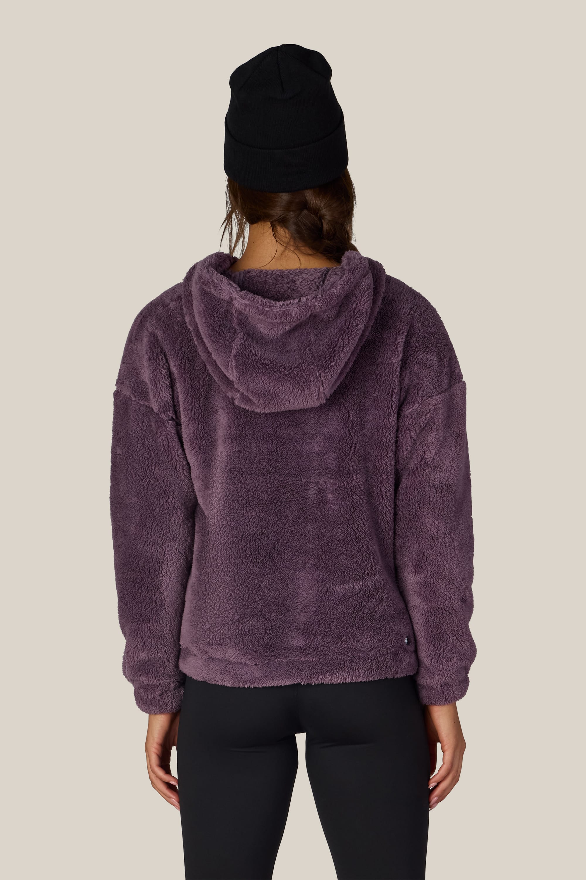 A person wearing a black beanie, the 686 Women’s Sherpa Hoody in purple, and black leggings stands facing away against a plain light background.