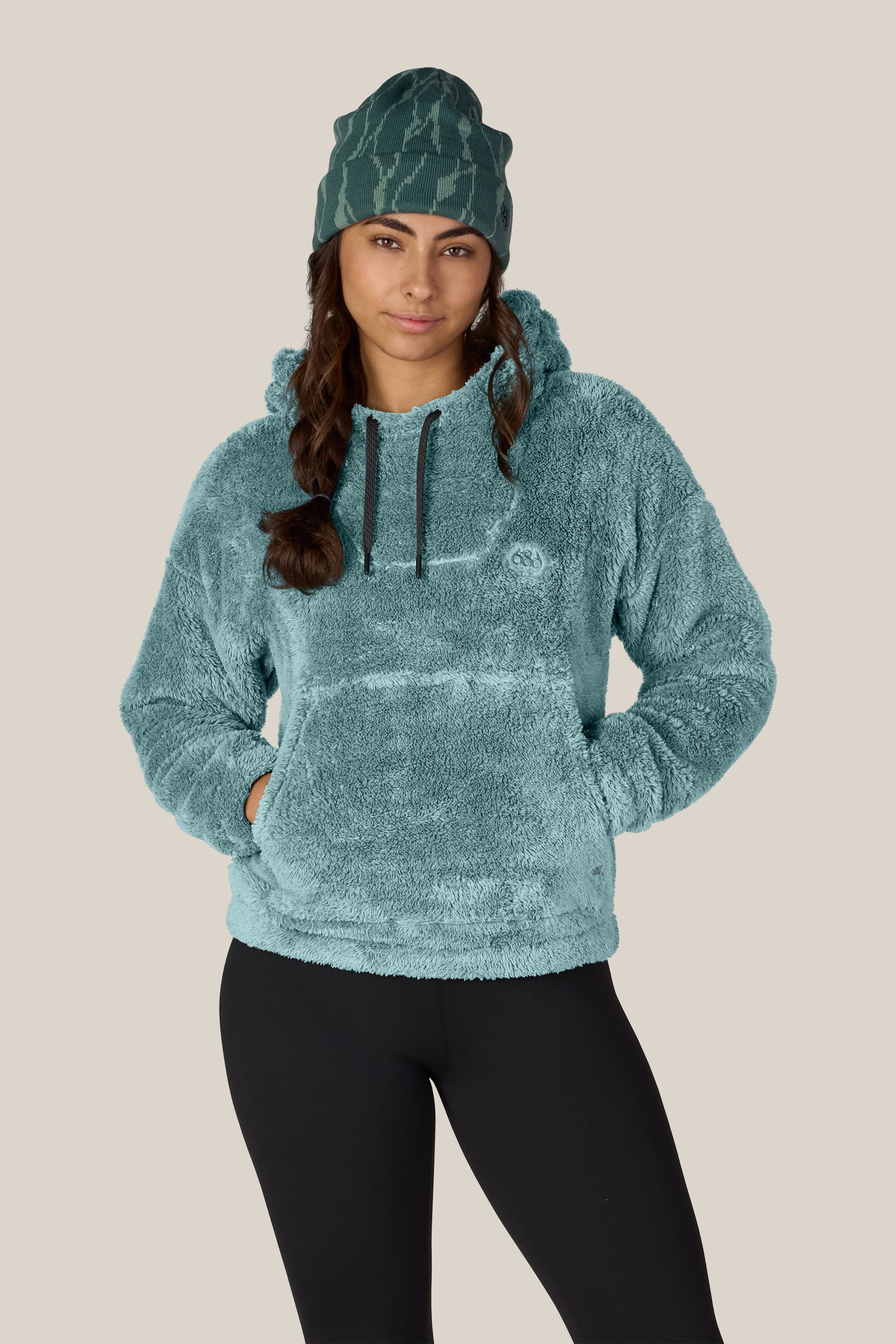A woman stands with her hands in the pockets of a 686 Women’s Sherpa Hoody by 686, paired with a teal patterned beanie and black leggings, against a plain beige background.