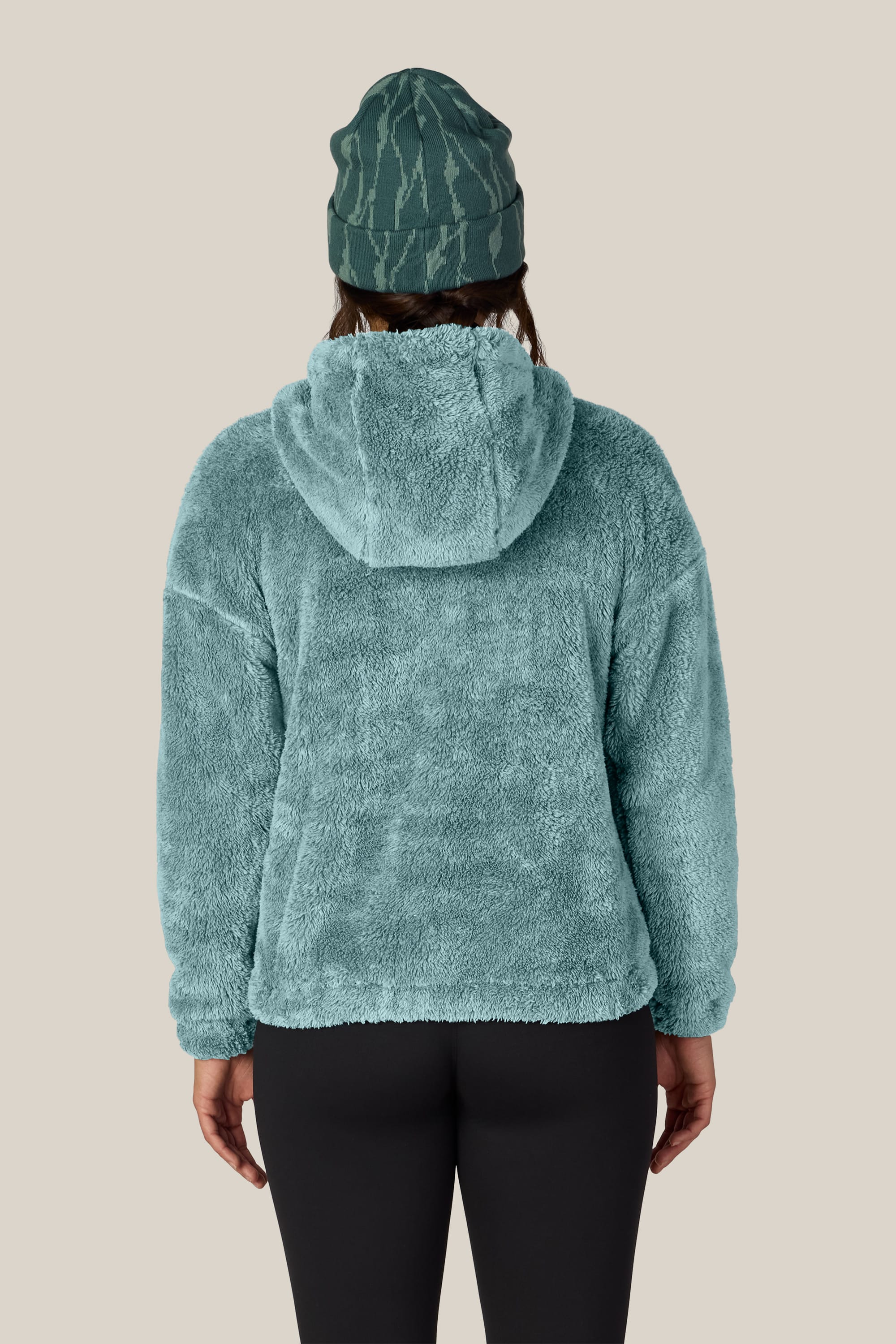 A person shown from the back against a plain light beige background wears the 686 Women’s Sherpa Hoody in teal, paired with black leggings and a green patterned beanie.
