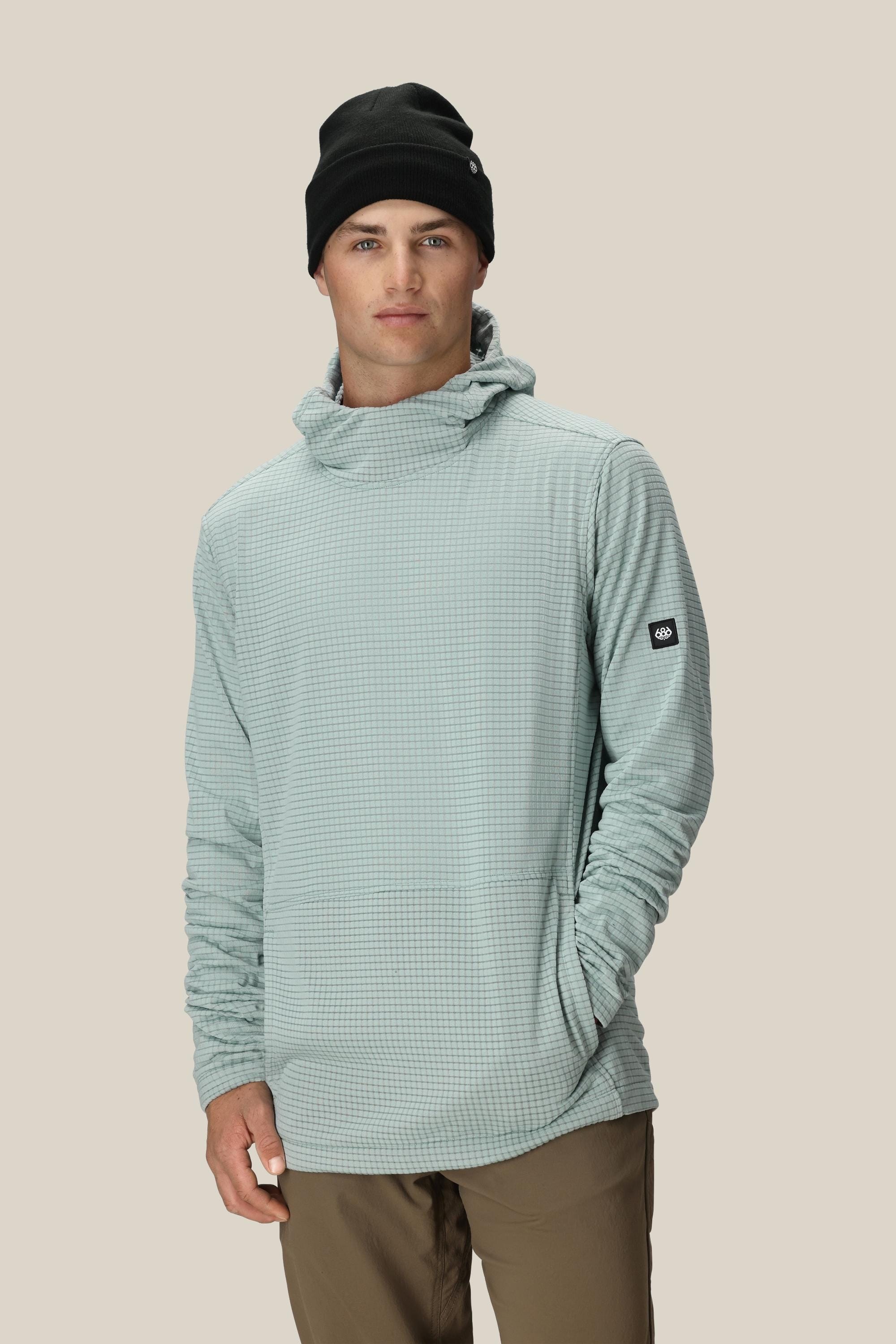 A man wearing the 686 Mens Mission Grid Fleece Hoody in light blue, brown pants, and a black beanie stands with one hand in his pocket, looking at the camera against a plain beige background.