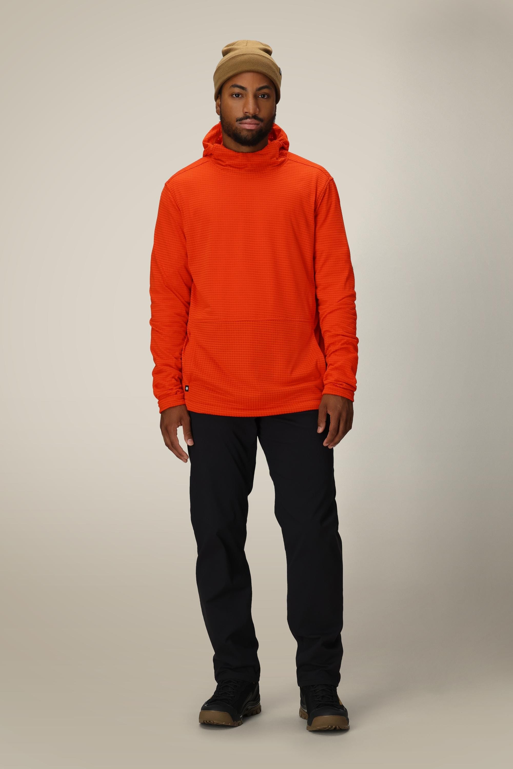 A man stands facing forward, wearing a tan beanie, the 686 Mens Mission Grid Fleece Hoody in orange, black pants, and brown shoes against a plain light background.