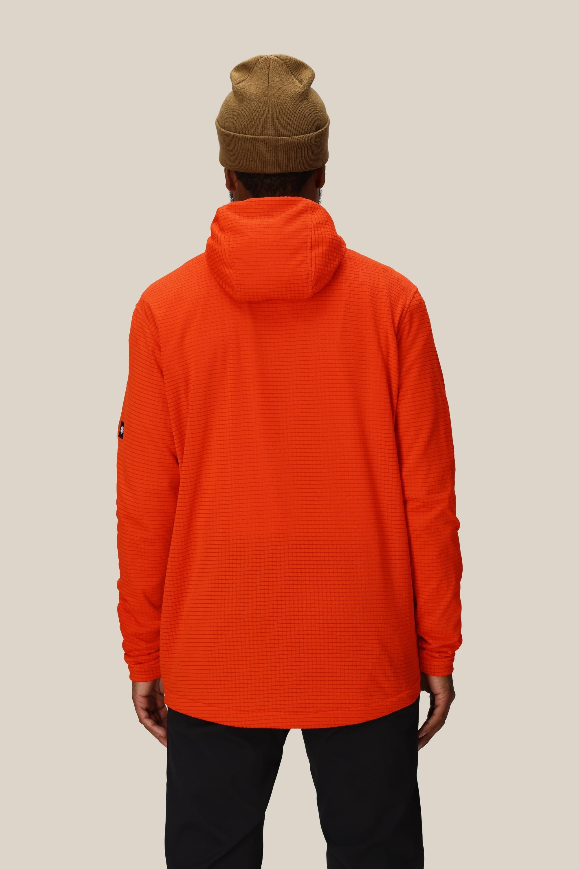 A person, seen from the back, wears the 686 Mens Mission Grid Fleece Hoody by 686 in bright orange and black pants, standing against a plain beige background.