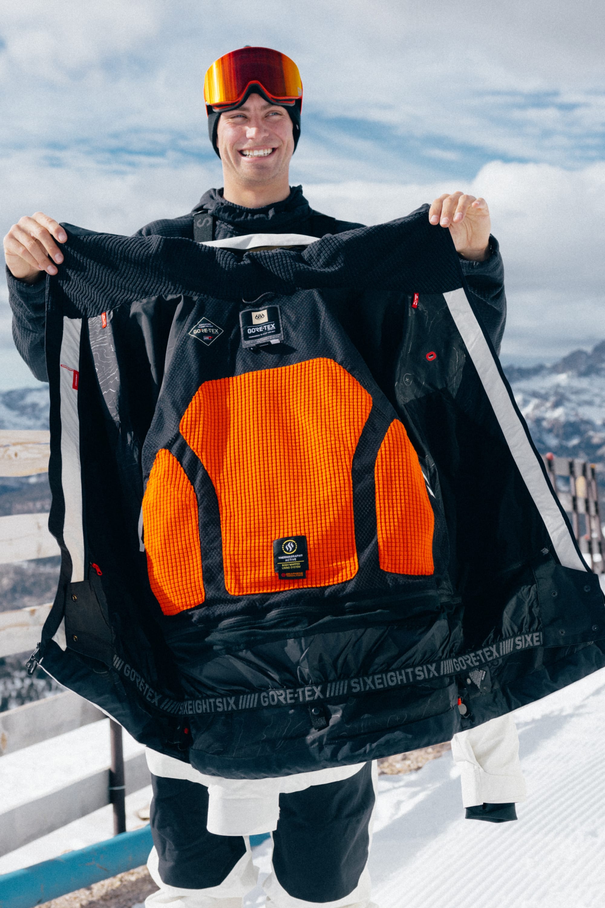 Wearing the 686 Men’s GORE-TEX® Dispatch™ Thermagraph® Jacket, a person dressed in ski gear and orange goggles stands on a snowy mountain, smiling as they hold open the black jacket to reveal its vibrant orange inner lining. Snowy peaks and a blue sky create a stunning backdrop.