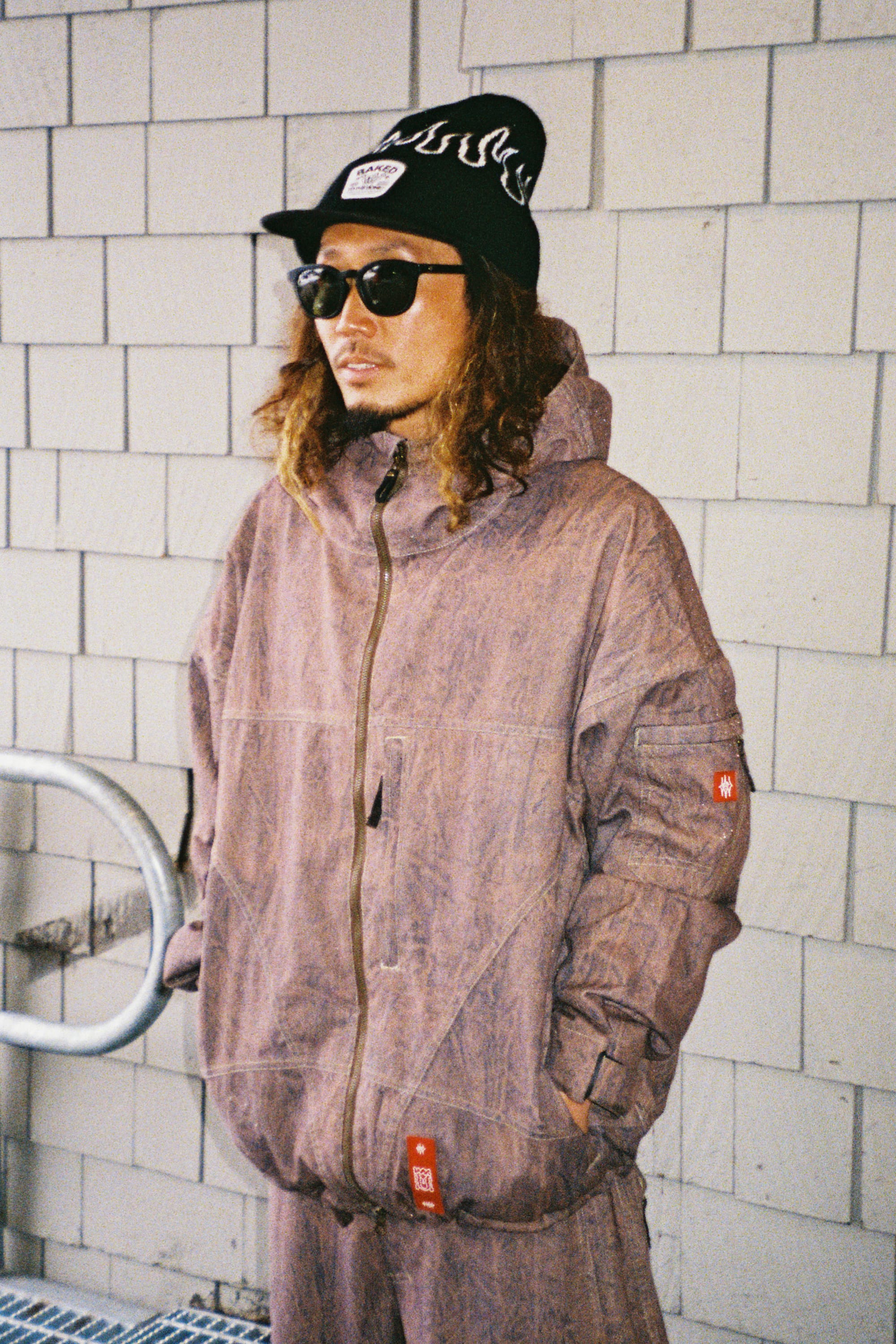 A person wearing the 686 Men’s 686 Dojo® Shell Jacket in purple, paired with black sunglasses and a black beanie featuring white flame designs, stands against a tiled wall with hands in pockets, gazing to the side.