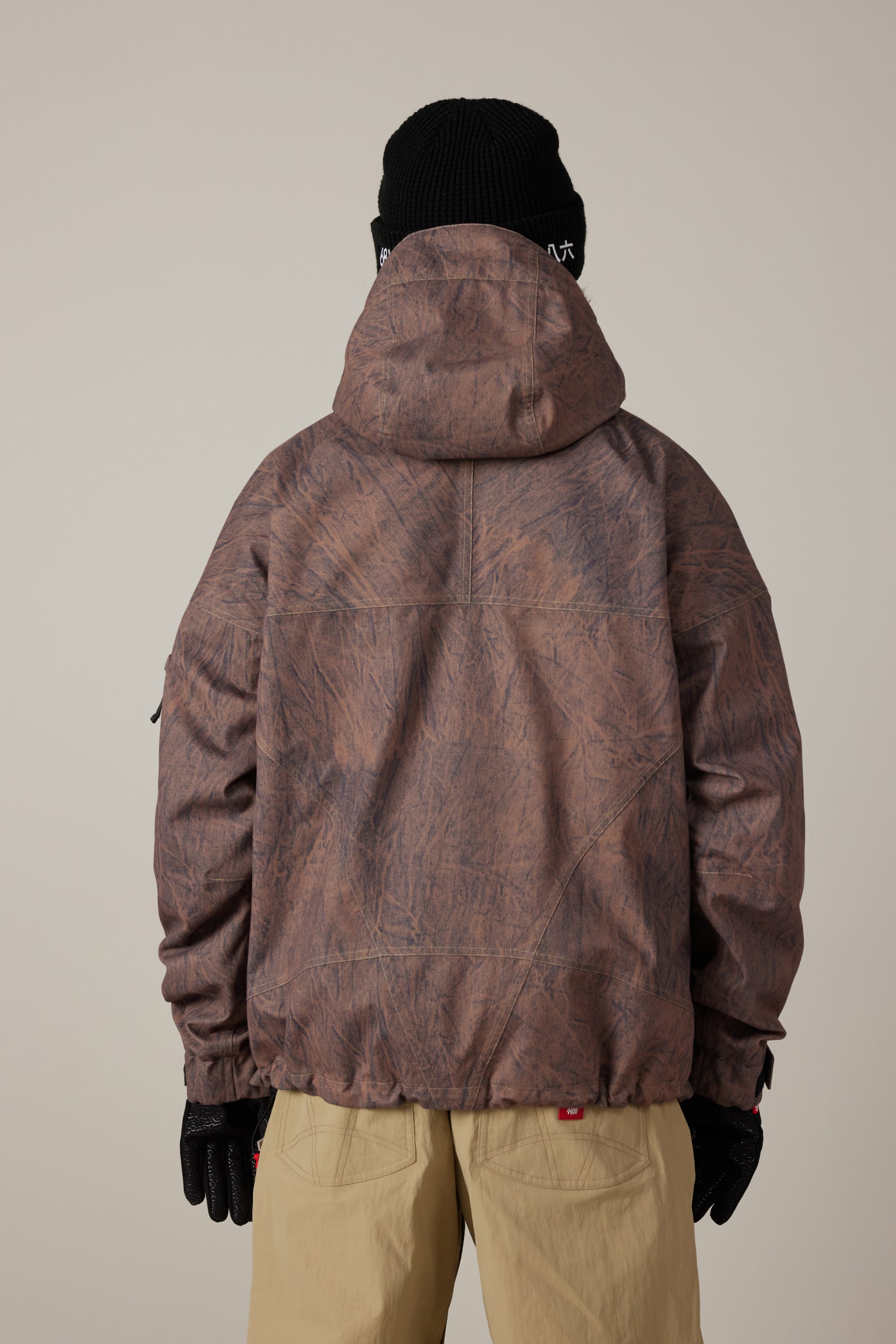 A person wearing the 686 Men’s 686 Dojo® Shell Jacket by 686—a brown, textured woven jacket with a hood—paired with a black knit beanie, black gloves, and beige pants, stands with their back to the camera against a neutral background.