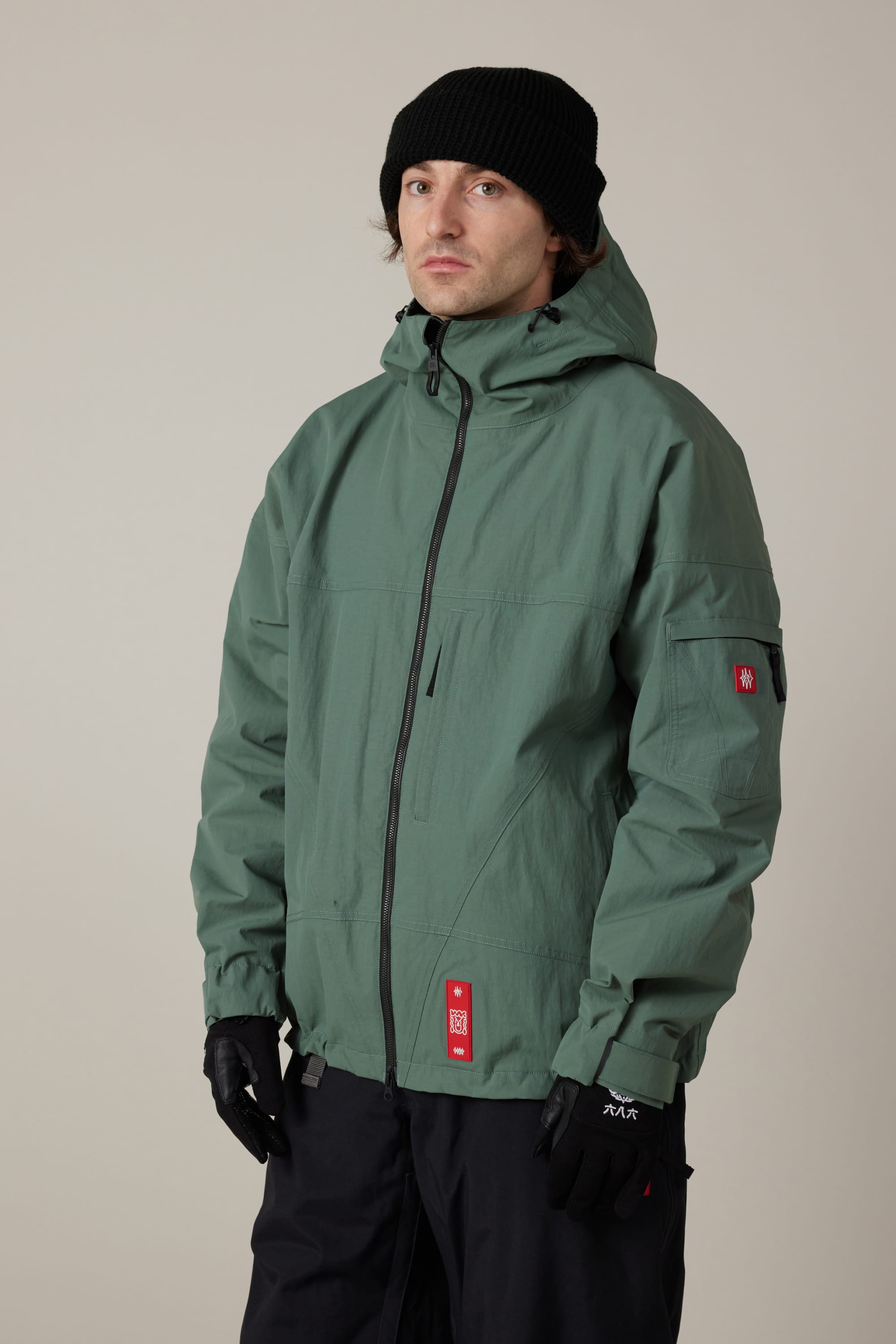 A person wearing the 686 Men’s 686 Dojo® Shell Jacket in green, paired with black gloves, a black beanie, and black pants, stands facing forward against a plain, light background.