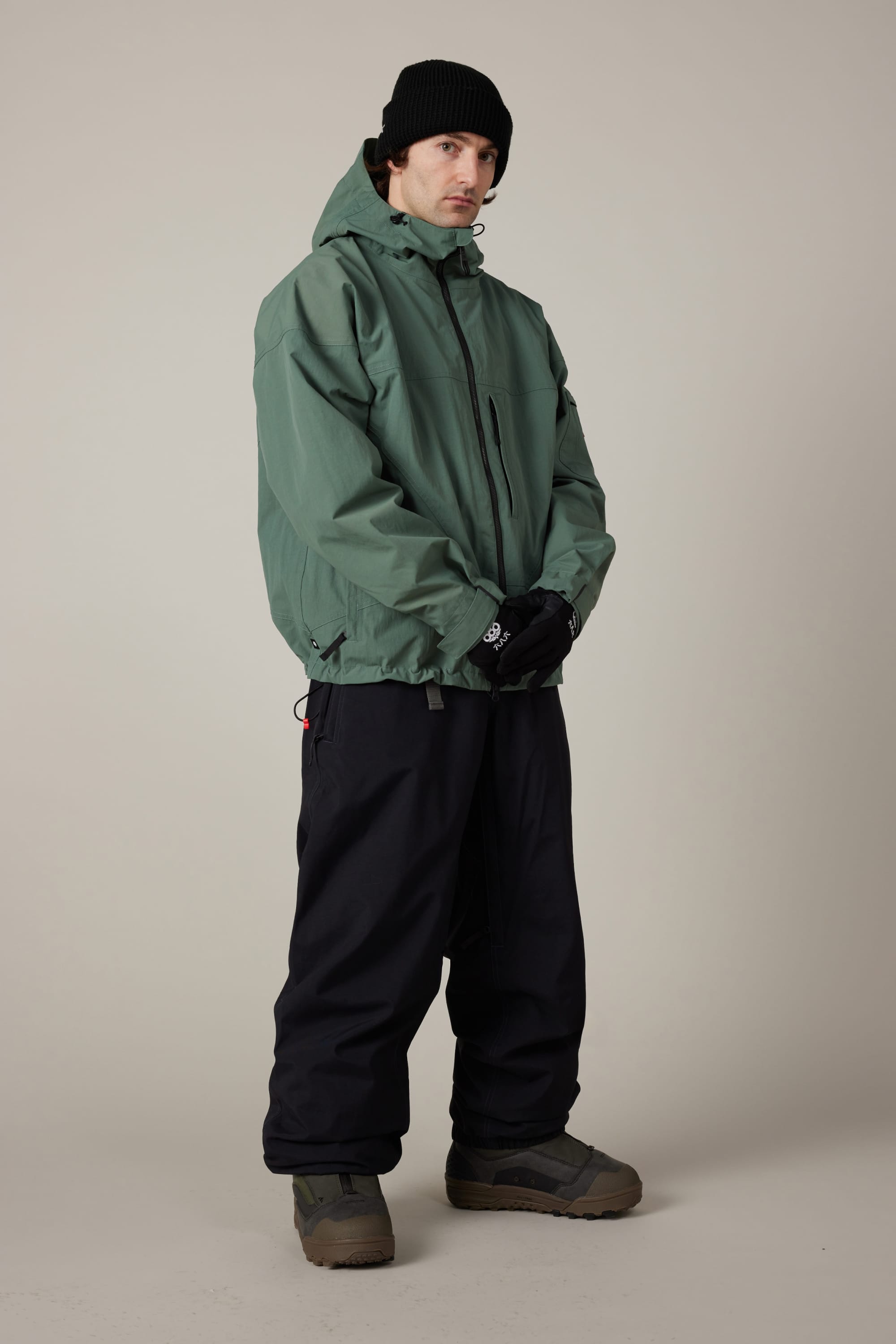 A person wearing the 686 Men’s 686 Dojo® Shell Jacket in green with a hood, paired with a black beanie, black gloves, loose black pants, and brown shoes stands against a plain light background, looking slightly to the side with hands clasped in front.