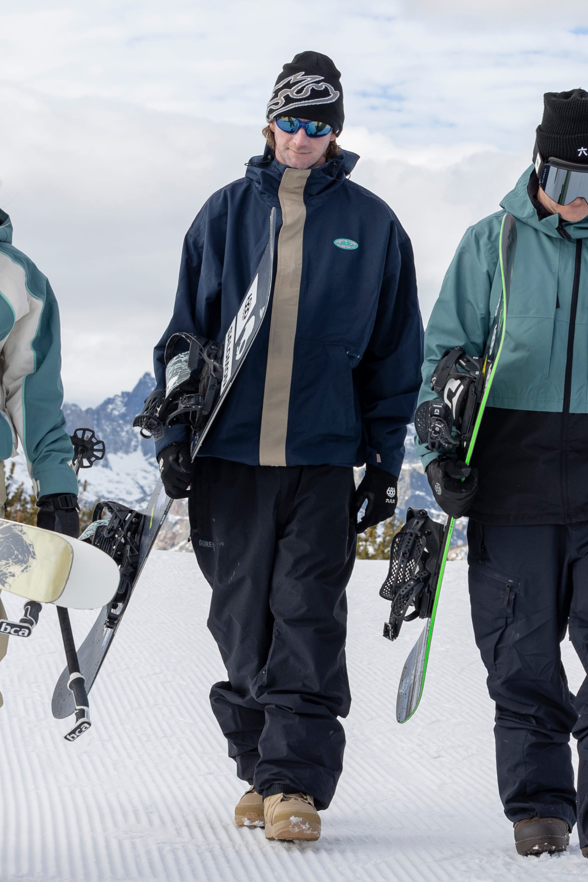 Three people in winter gear walk on snow, carrying snowboards. The central person is wearing the 686 Men’s 1994 Liberty Jacket, a beanie, blue sunglasses, and tan boots. Snowy mountains and a cloudy sky are visible in the background.