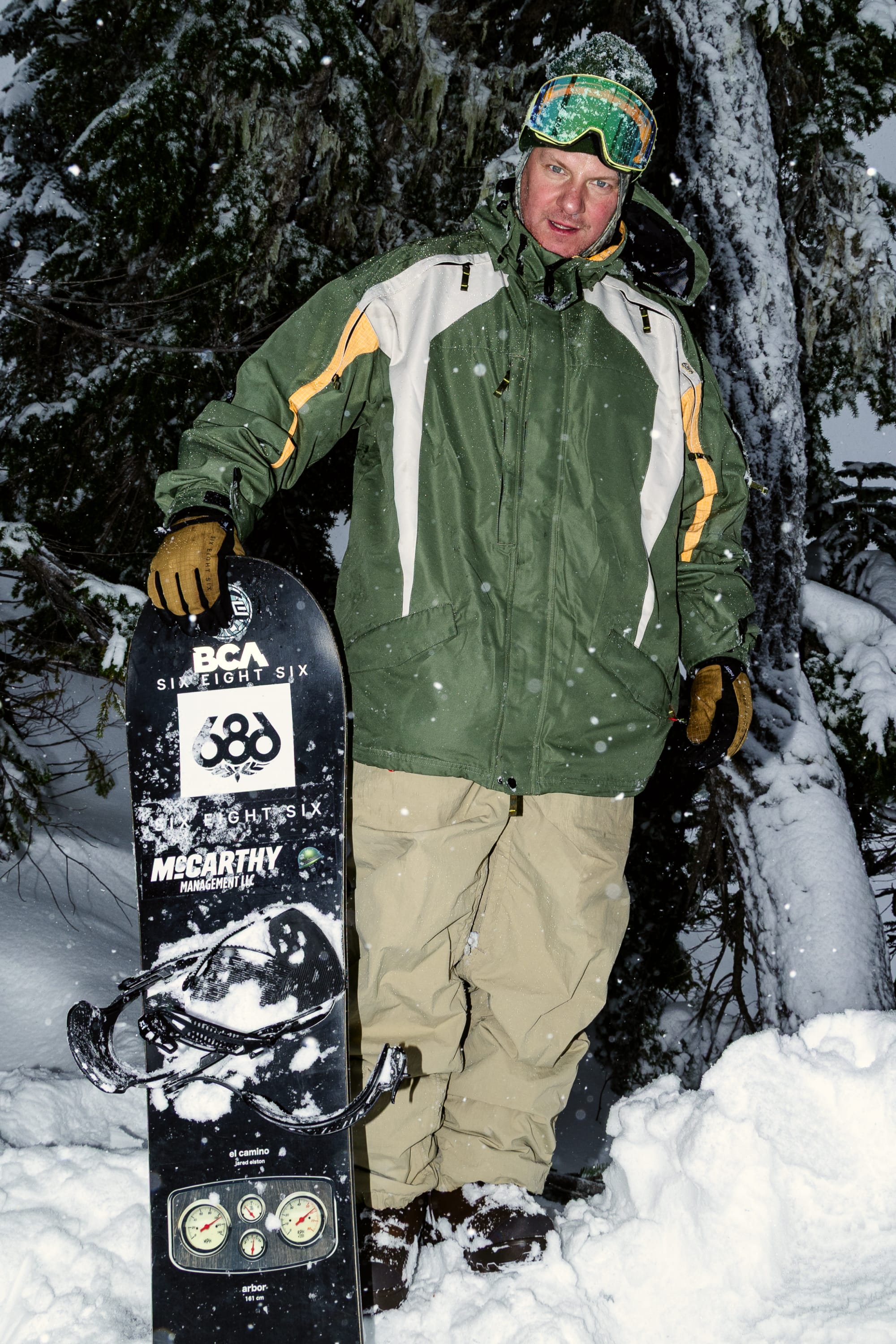 Wearing the 686 Men’s 2003 Atom Jacket, a person in green and tan pants stands in the snow holding an upright snowboard dusted with snow, surrounded by snow-covered trees. They are also wearing goggles and gloves.